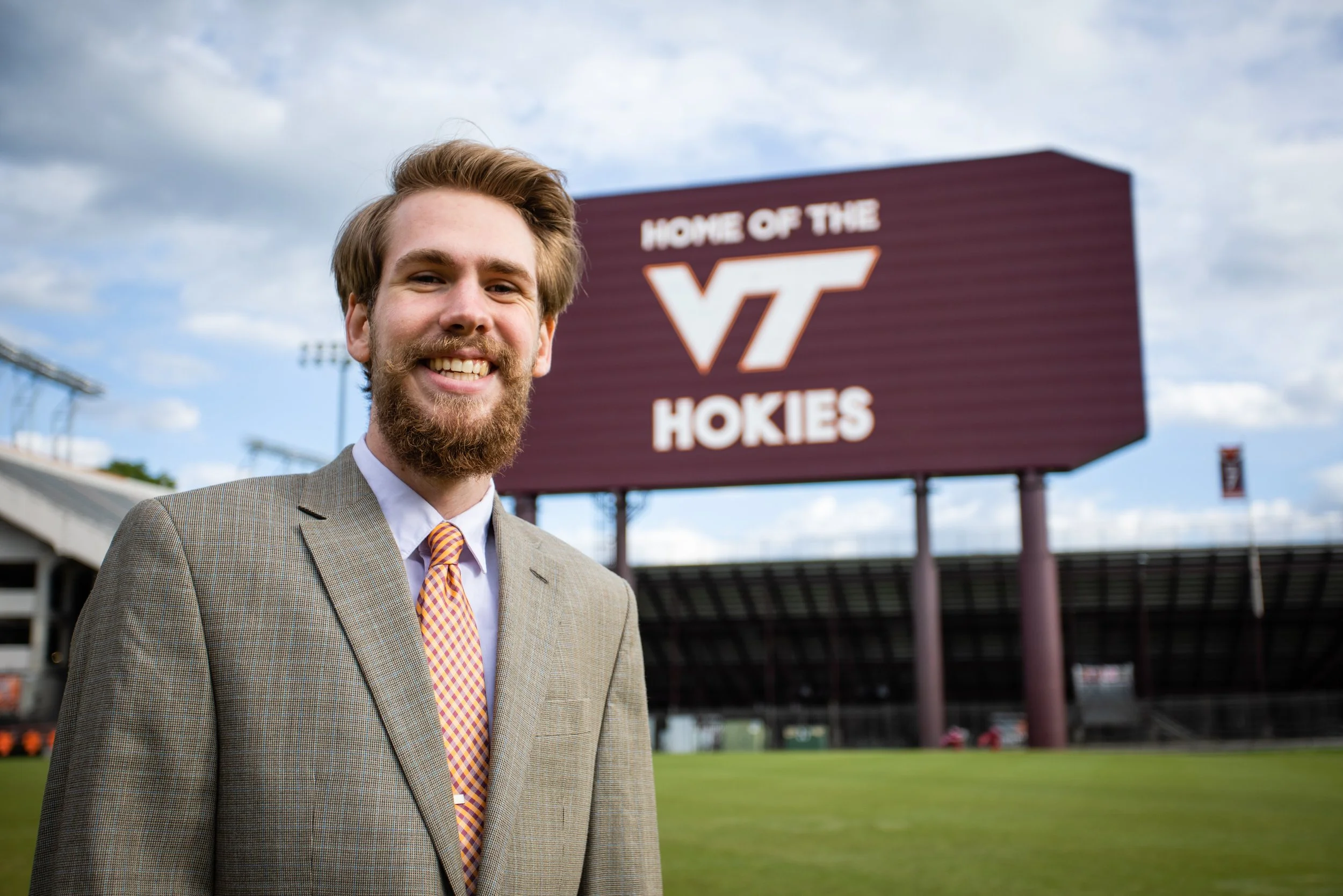 A man in a suit standing on a football field with a large sign in the background that reads 'Home of the VT Hokies'.