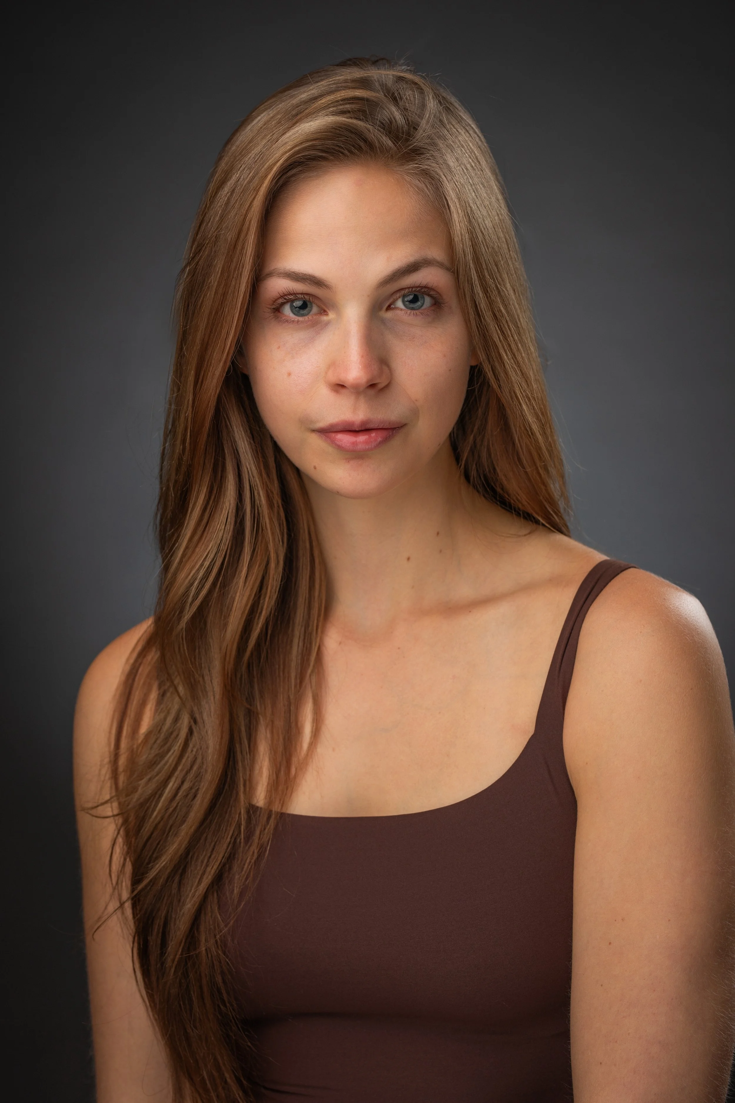 A young woman with long, wavy brown hair and blue eyes, wearing a brown tank top, looking directly at the camera with a slight smile against a dark gray background.