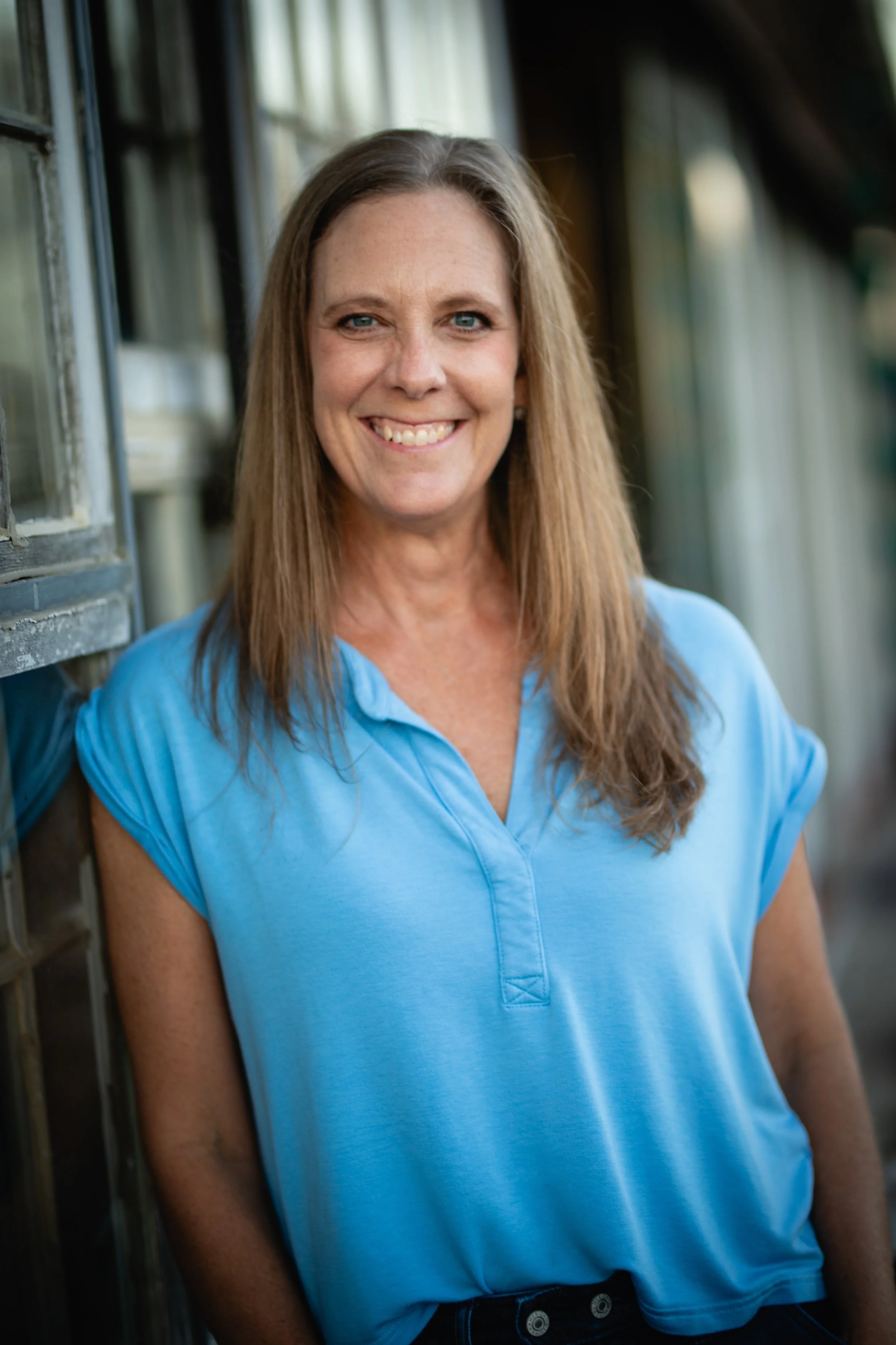 A woman with long light brown hair and a bright smile, wearing a light blue short-sleeved shirt, leaning against a rustic wooden window frame outside.