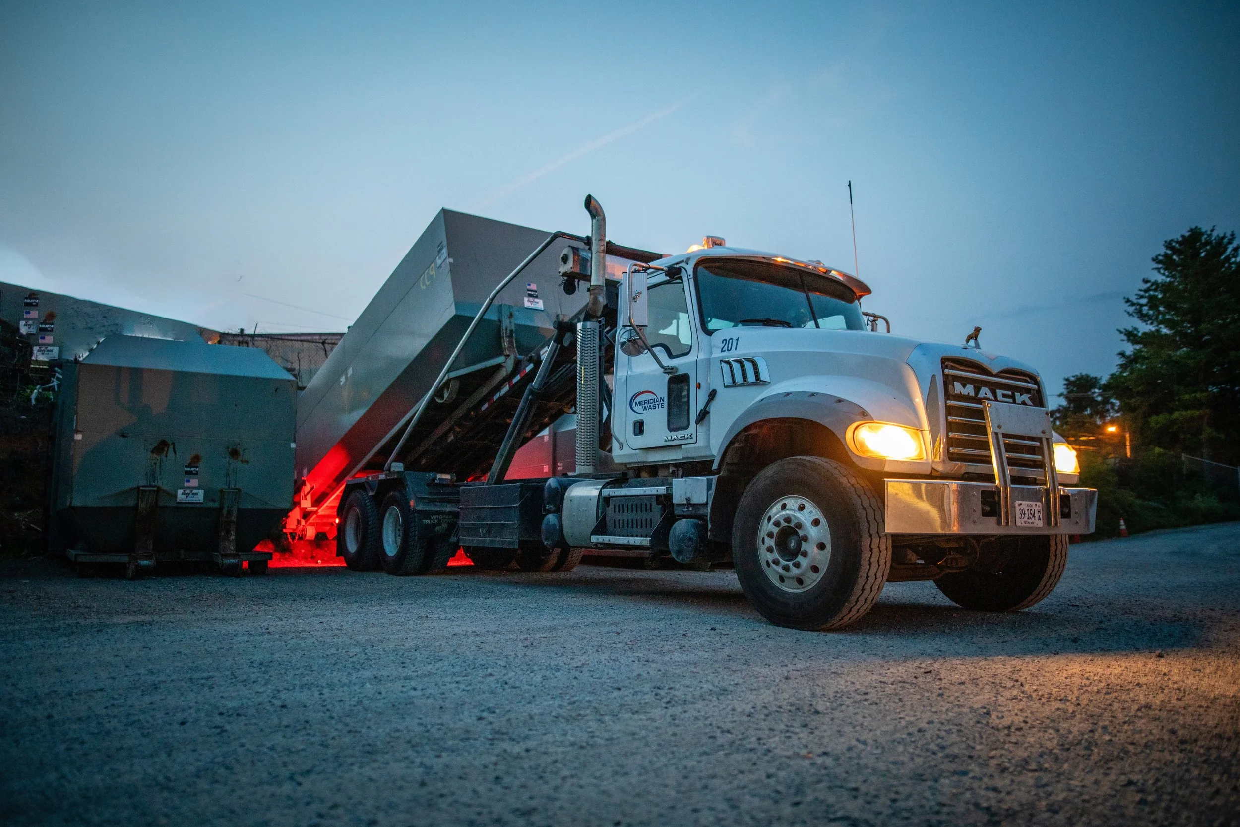 A white Mack waste collection truck in a gravel lot during dusk, collecting trash containers with red lights illuminating the area.
