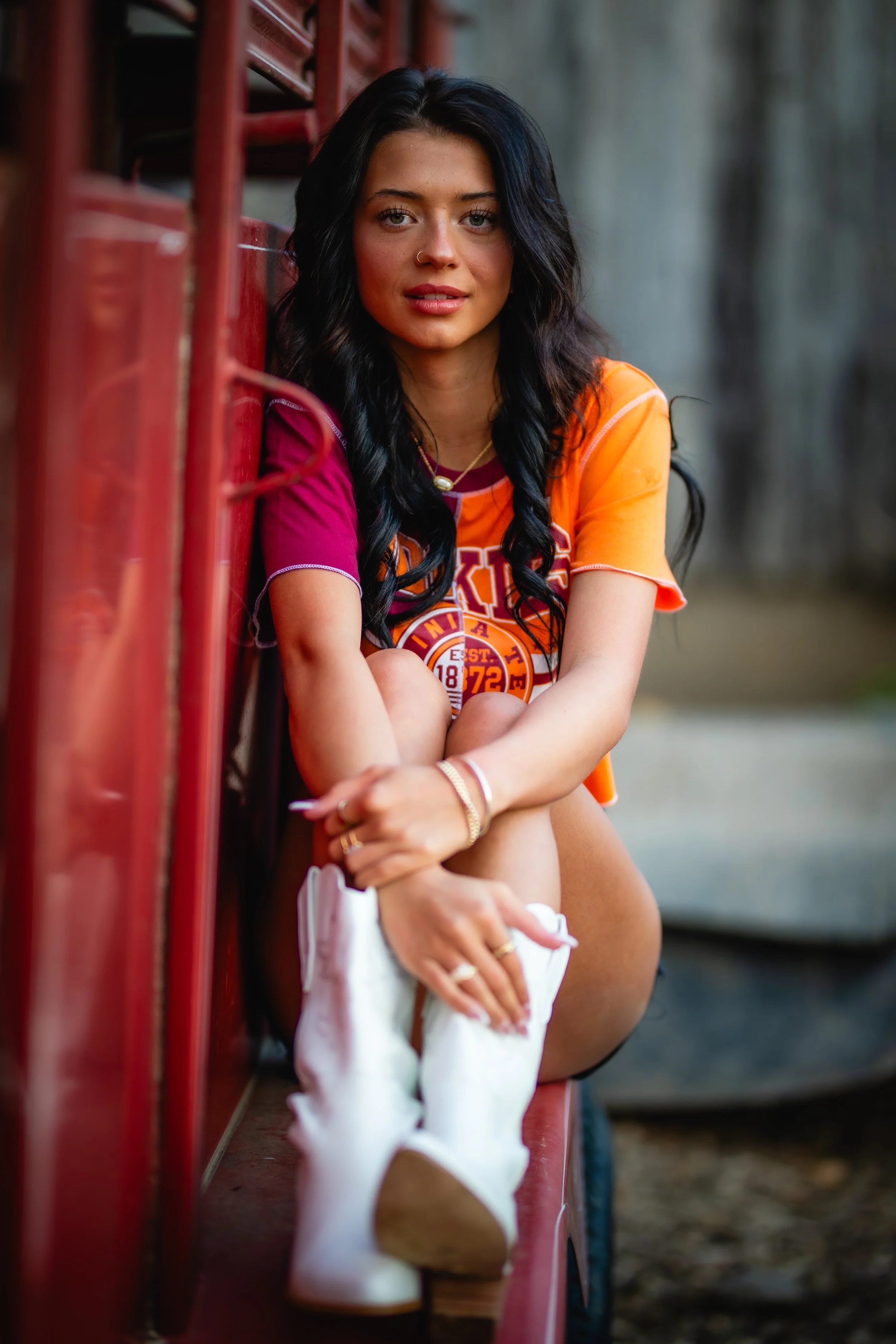 A young woman with black hair, sitting on a red vehicle, wearing a colorful sports jersey, white boots, and jewelry.