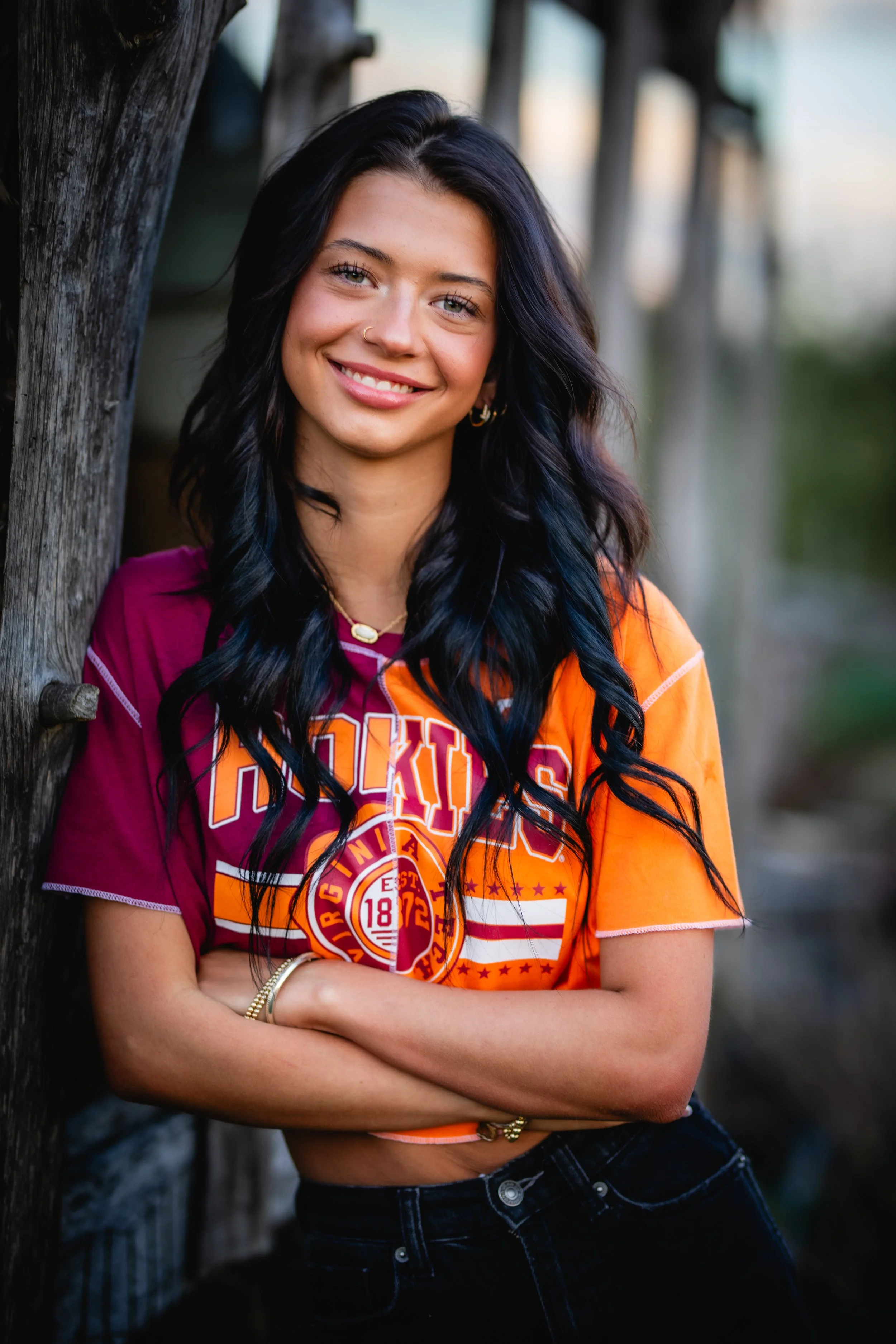 A young woman with long dark wavy hair, wearing a colorful college sports shirt with orange, purple, and white colors, is leaning against a tree outdoors and smiling at the camera.