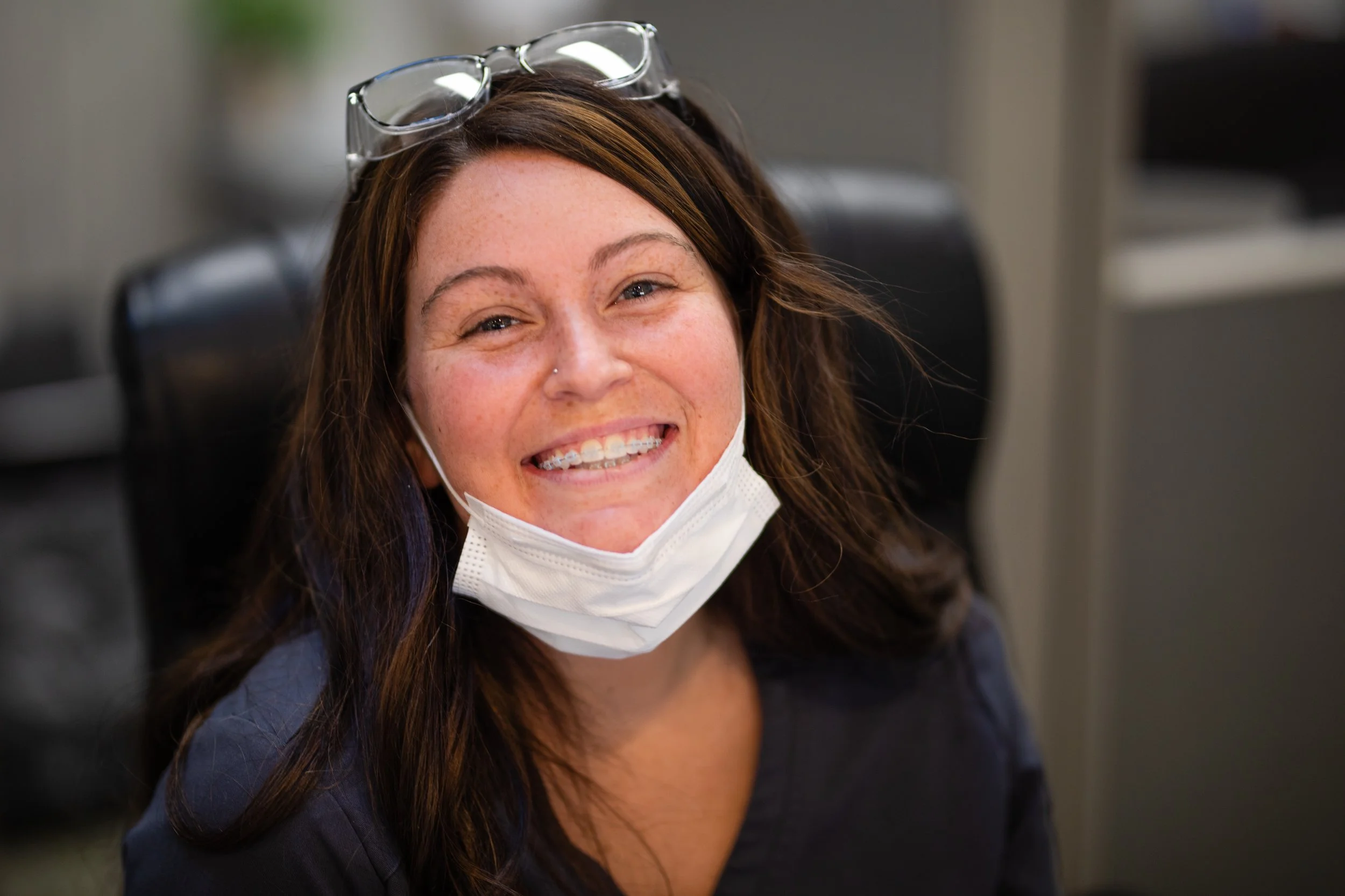 A woman with long brown hair smiling, wearing glasses on her head and a face mask pulled down, sitting in an office chair.