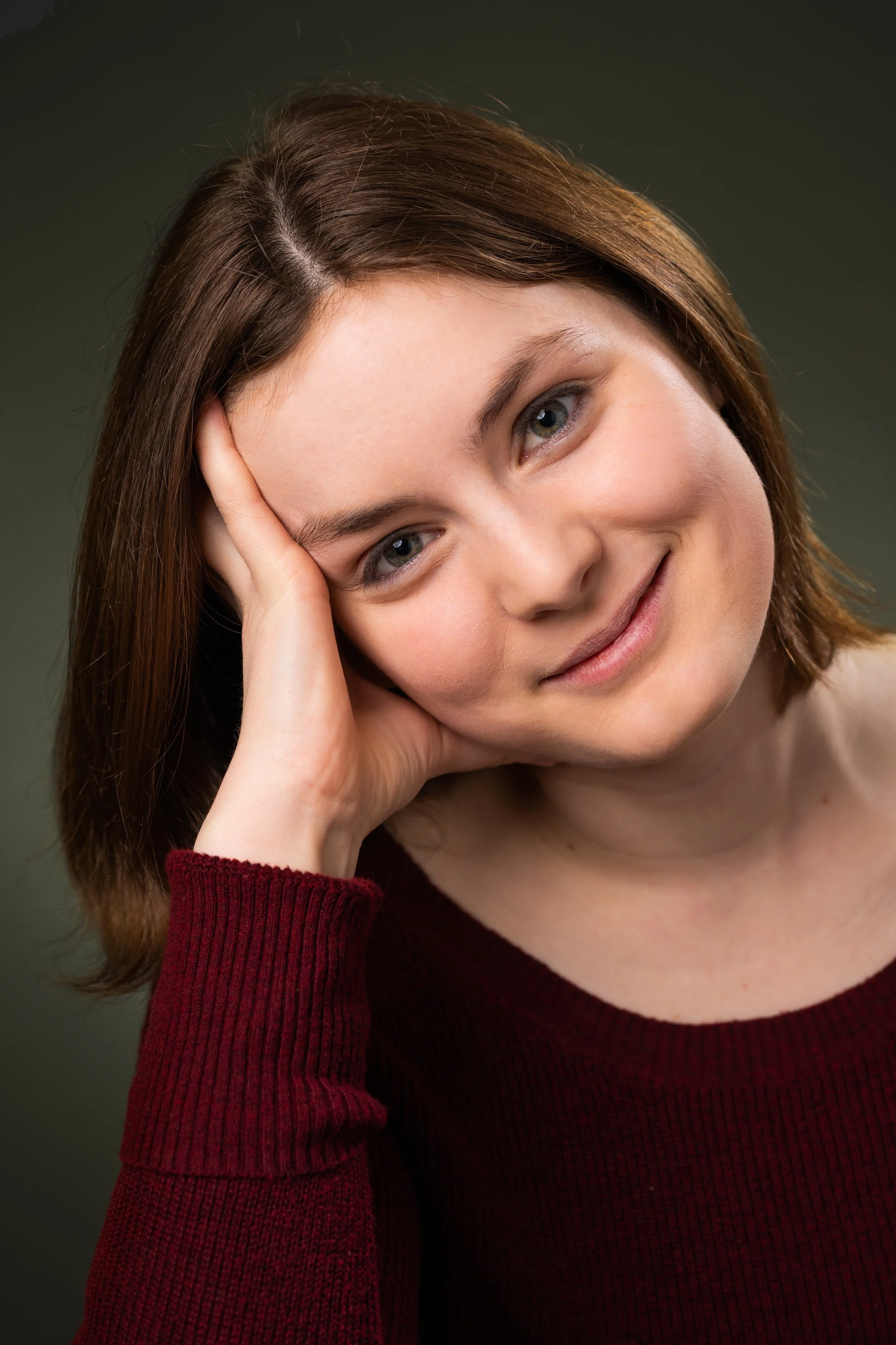 A woman with shoulder-length brown hair and blue eyes smiling gently, resting her head on her hand, wearing a red sweater, against a dark background.