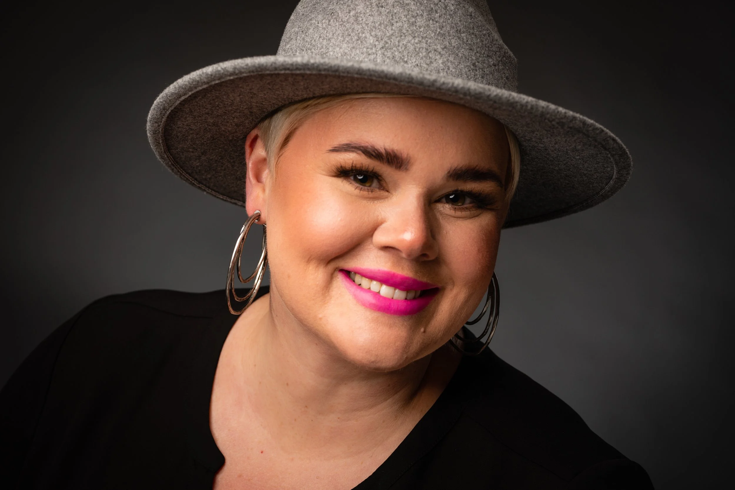 A woman with short blonde hair, wearing a gray fedora hat, pink lipstick, hoop earrings, and a black top, smiling against a dark background.