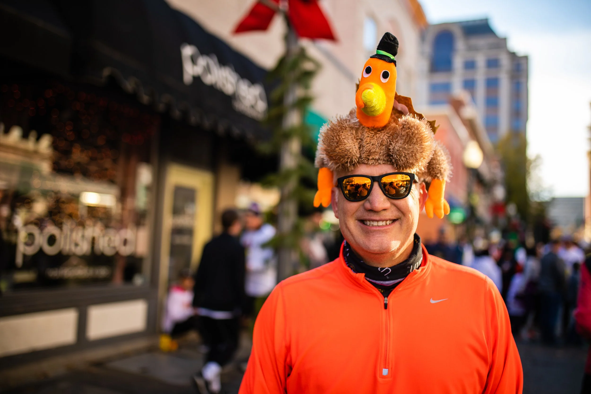 A smiling man wearing sunglasses and an orange running jacket stands outdoors in a busy urban area during daylight. He has a humorous hat with a plush turkey that has googly eyes and a small witch's hat on top.