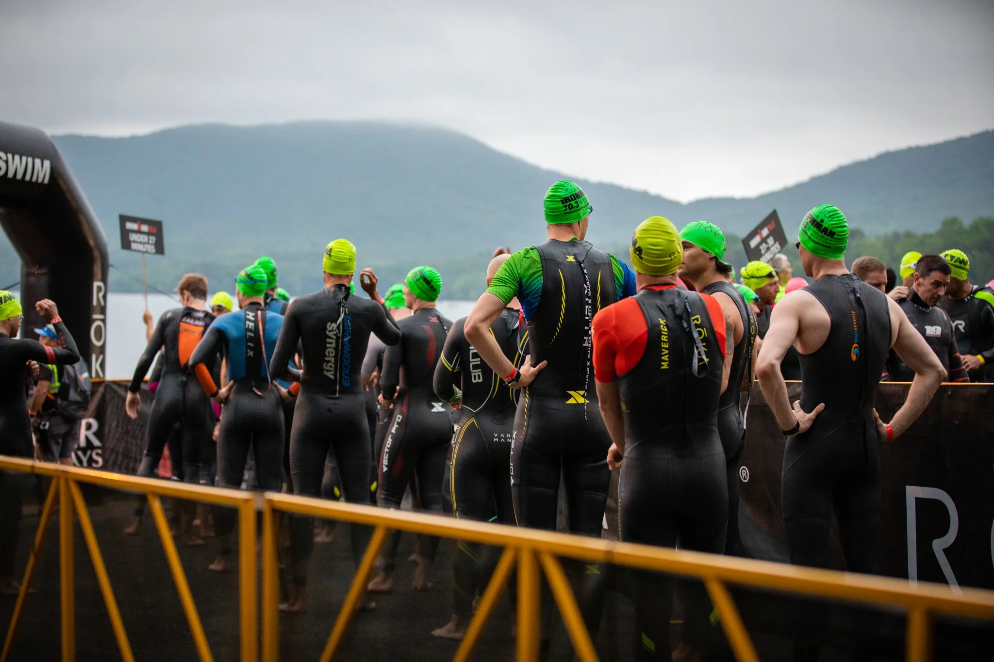 Triathletes in wetsuits and green swim caps gather at the starting area near the water with mountains in the background before a race.