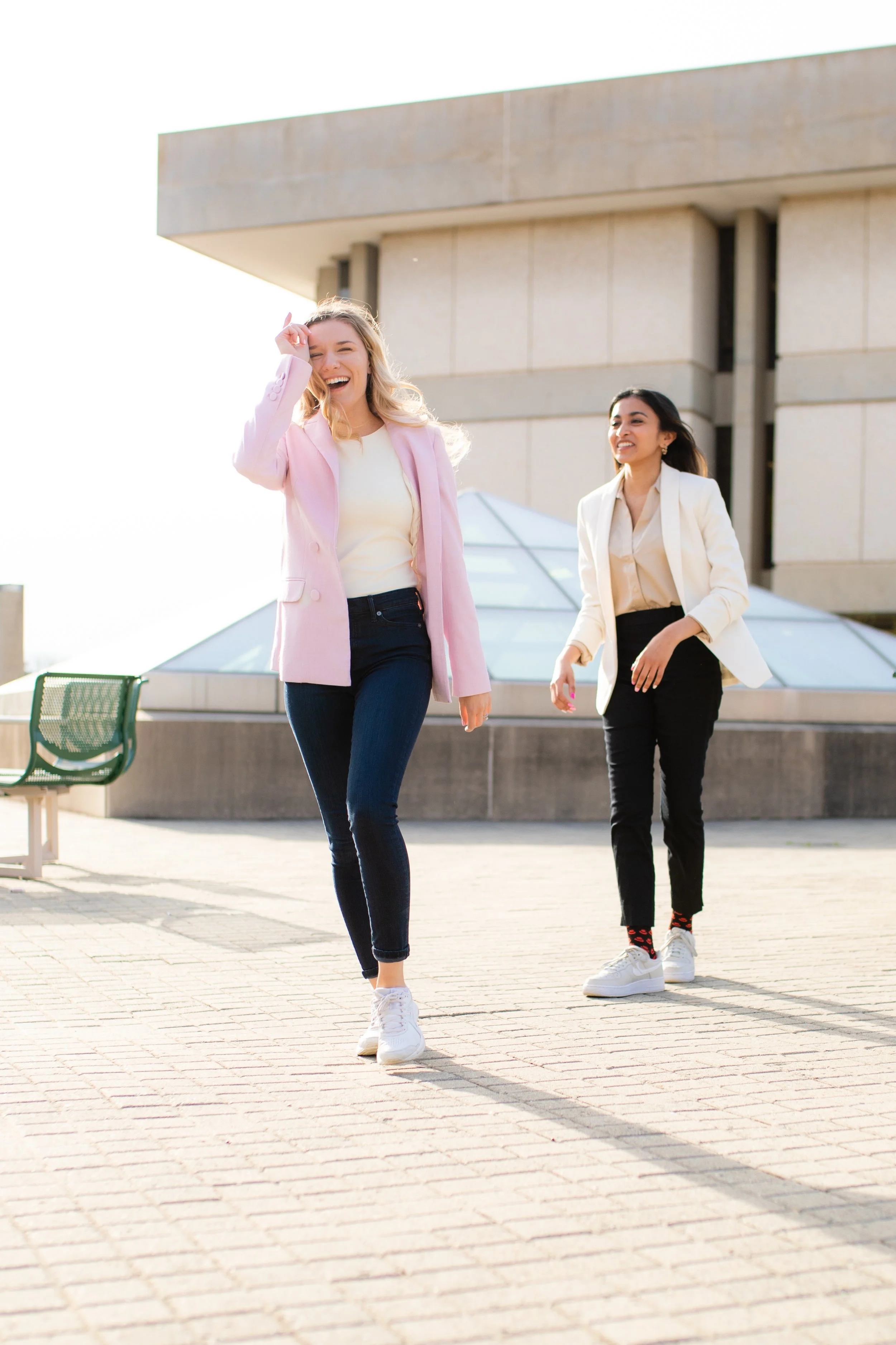 Two young women walking and smiling outdoors on a sunny day; one in a pink blazer and jeans, the other in a white blazer and black pants.