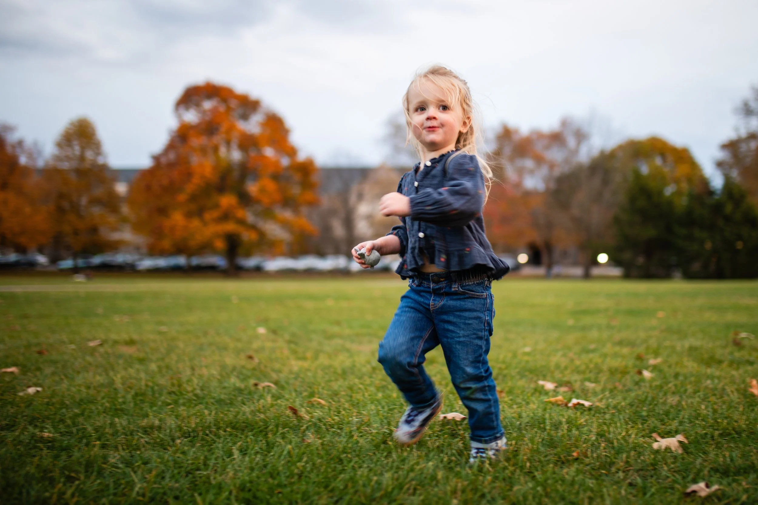 A young girl with blonde hair wearing a dark blue jacket and jeans running on a grassy field in autumn with trees in the background.