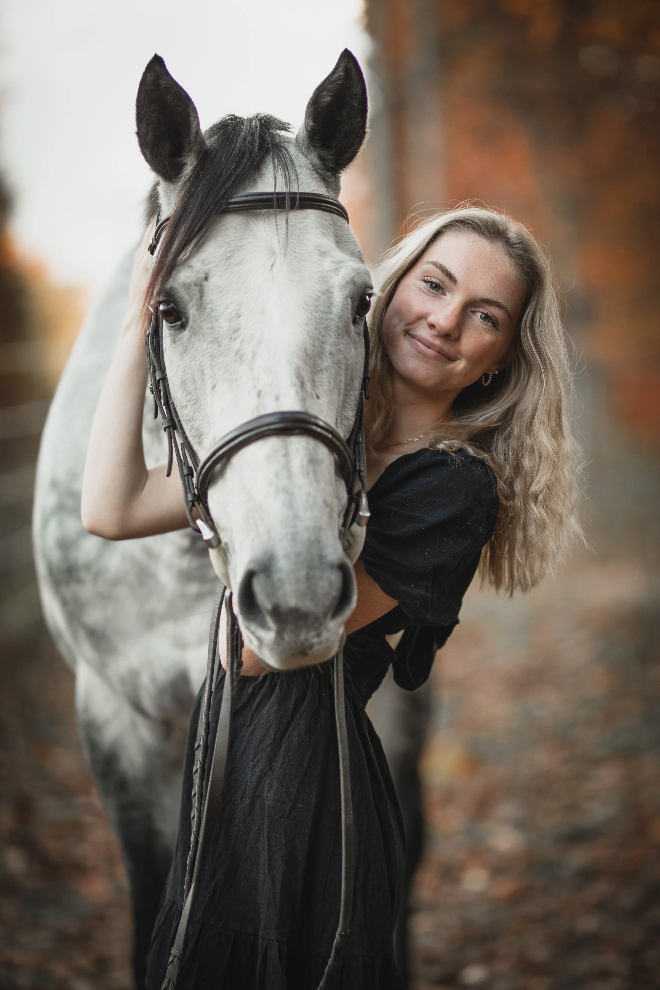 A young woman with wavy blonde hair and a black dress standing next to a white and grey horse outdoors during autumn.