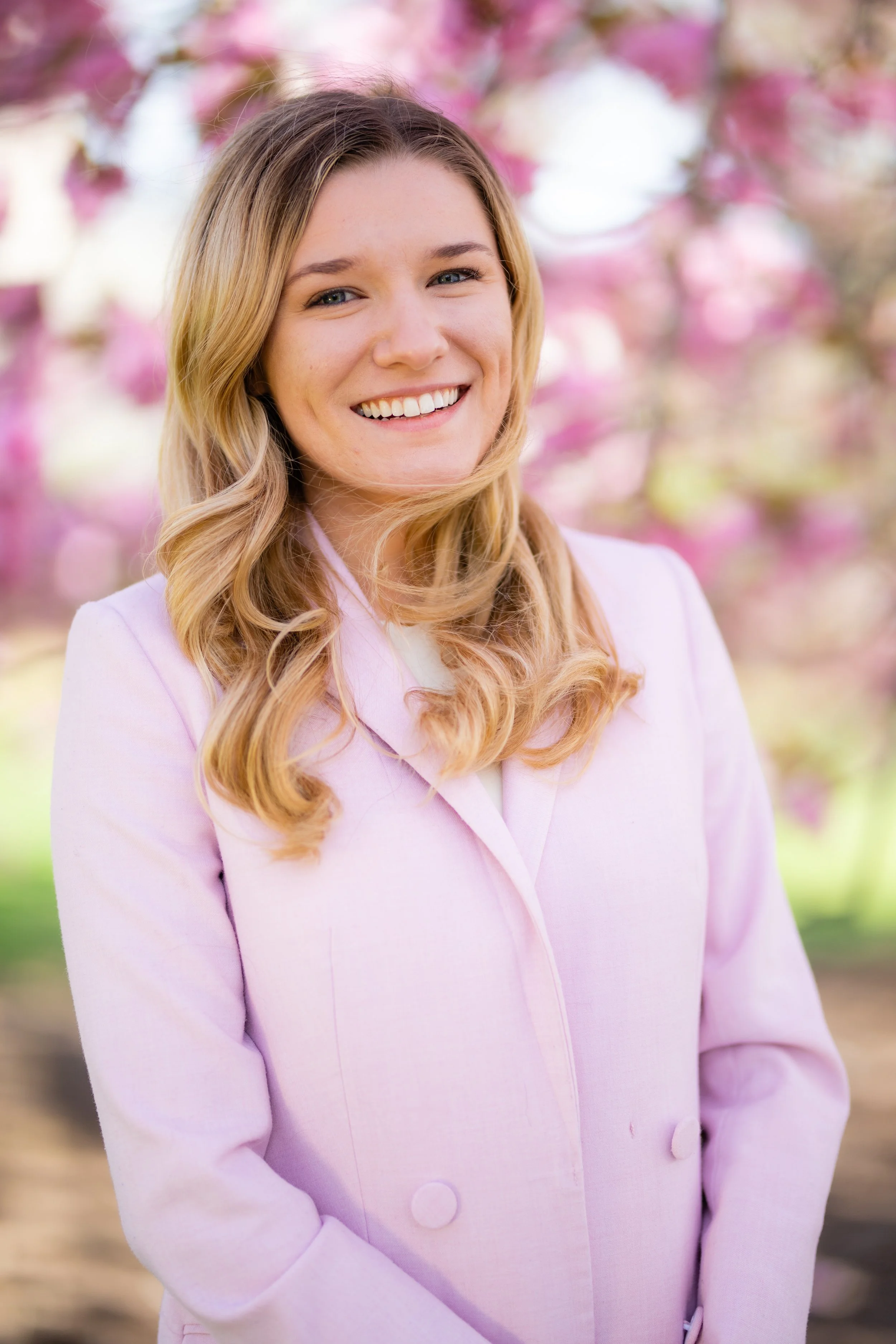 A smiling woman with long, wavy blonde hair wearing a light pink blazer, standing outdoors with pink flowering trees in the background.