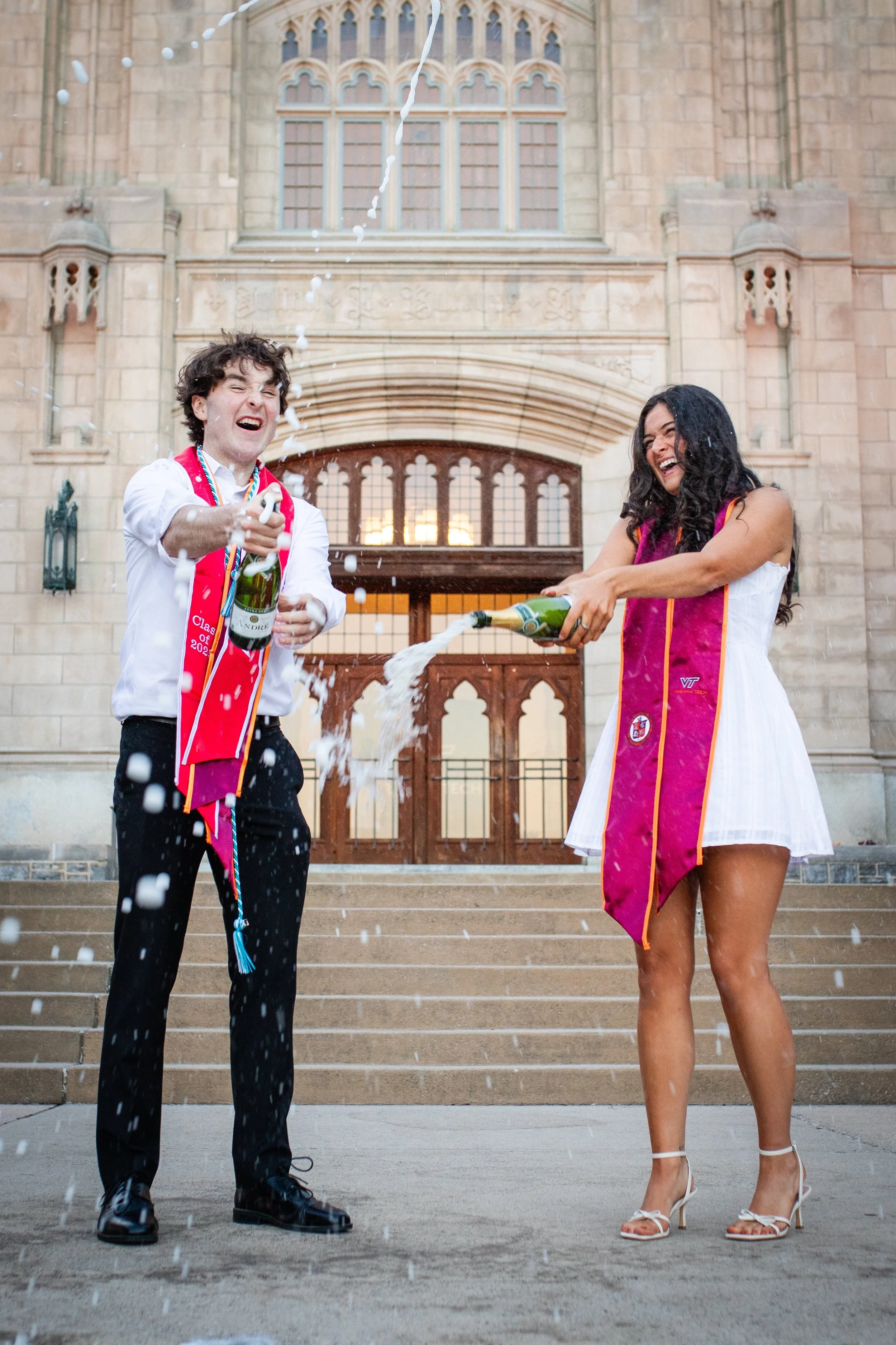 Two graduates, one man and one woman, celebrate with champagne outside a university building. They are smiling, wearing graduation stoles, and spraying champagne in the air.