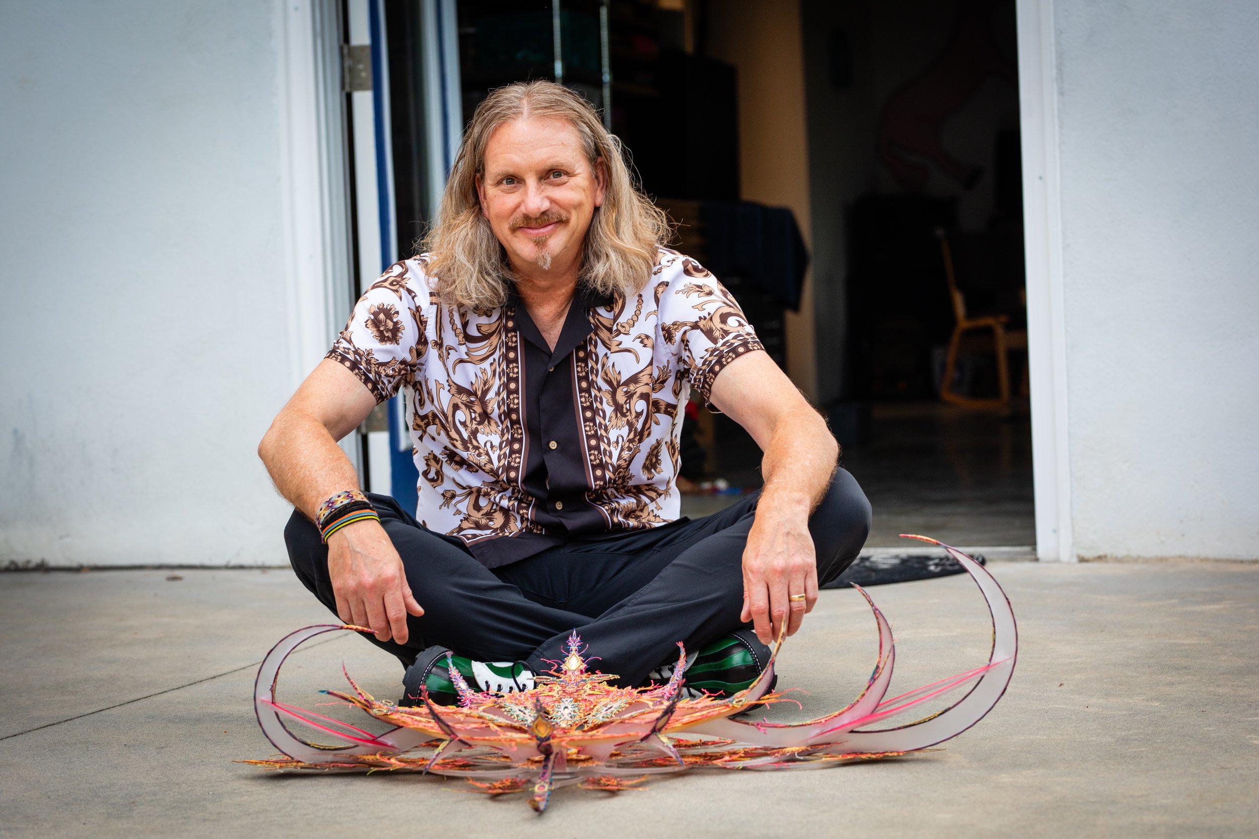 Man with long hair and goatee sitting cross-legged outside, smiling, next to a large colorful art sculpture on the ground.