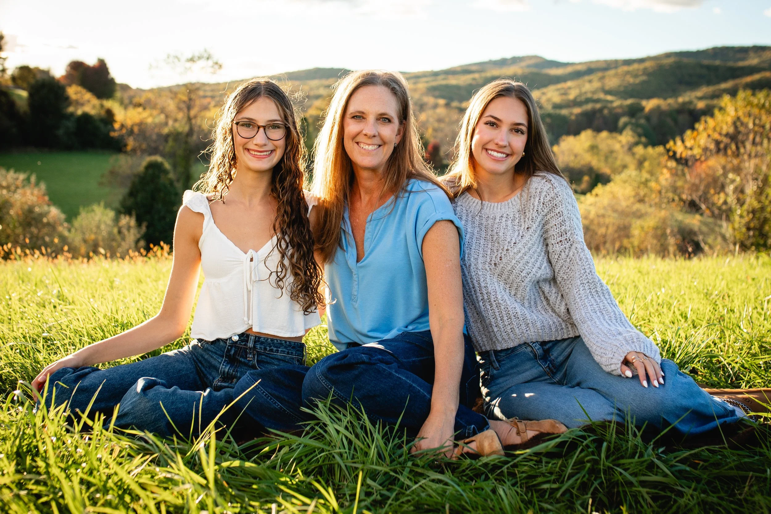 Three women sitting on grass in a field with trees and hills in the background during sunset, smiling at the camera.