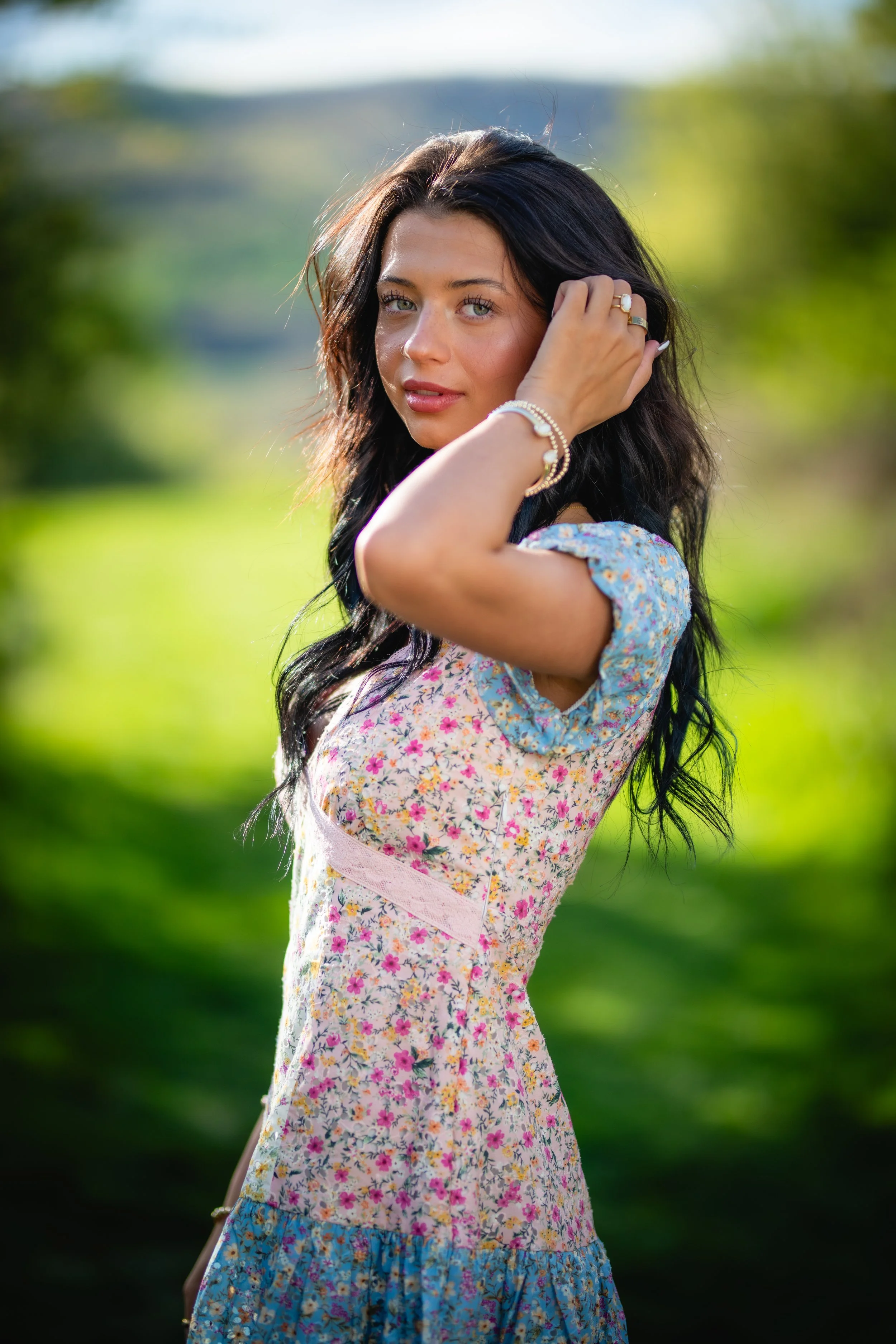 A young woman with black, wavy hair posing outdoors in sunlight, wearing a floral dress with short sleeves and jewelry, with a blurred green background.