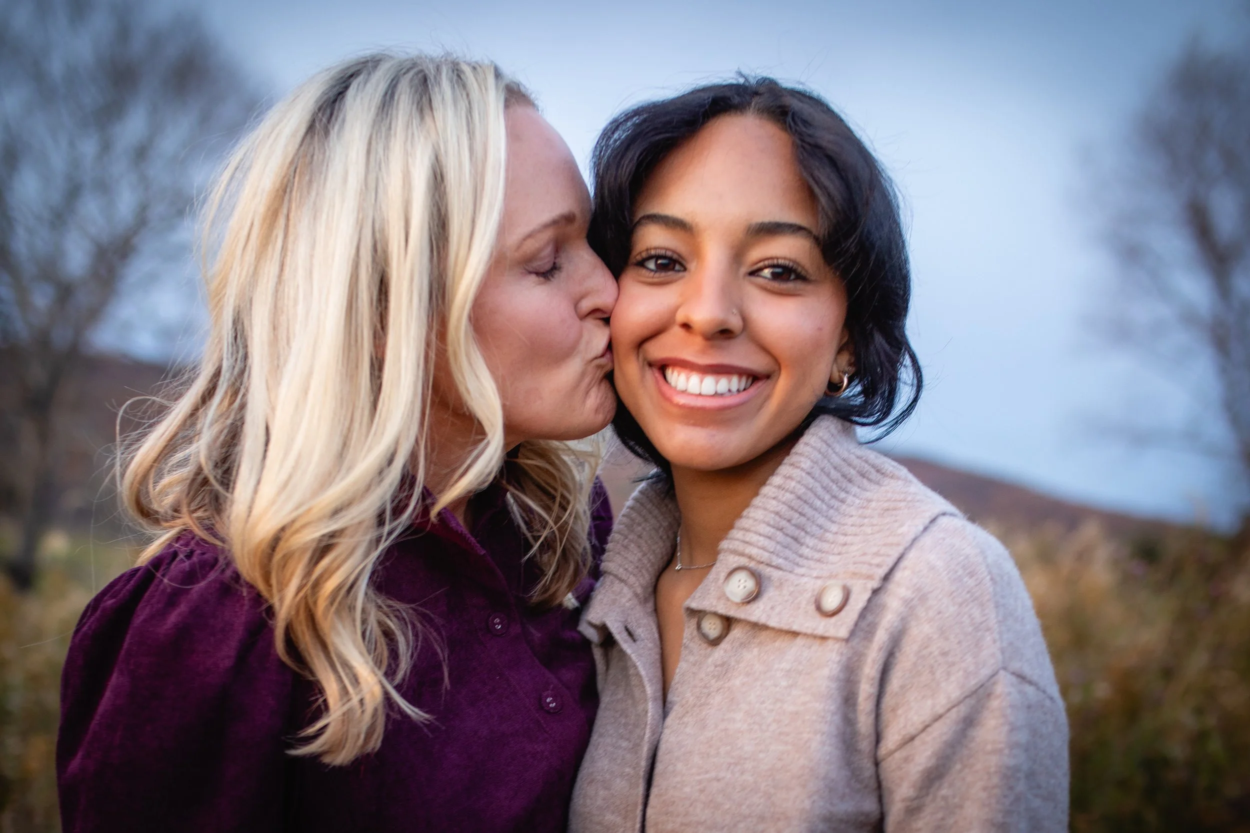 Two women, one with blonde hair and the other with short black hair, sharing a kiss on the cheek outdoors during daytime, with trees in the background.
