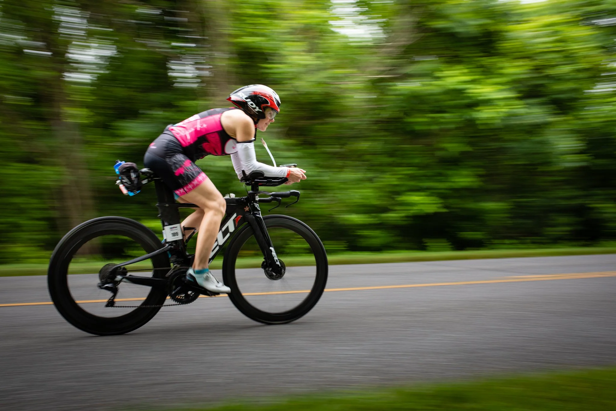 A person in cycling gear riding a black Trek bicycle on a road, with a blurred green forest background, during daytime.