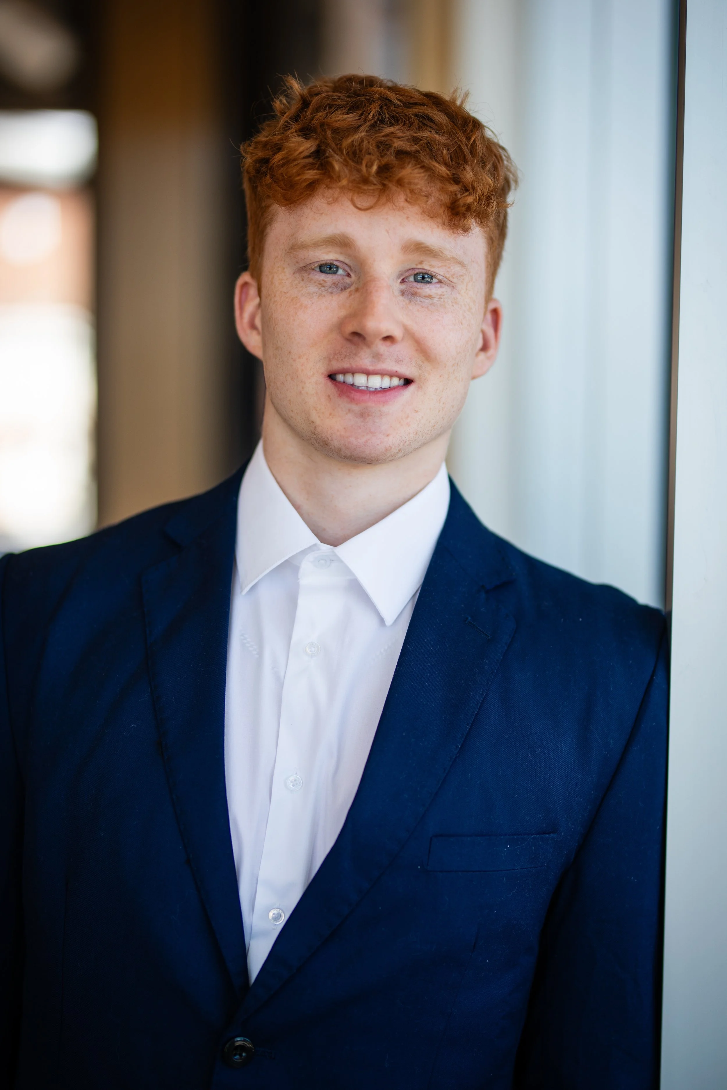 Young man with ginger hair, blue eyes, and freckles smiling, wearing a dark blue suit and white shirt, standing by a window.