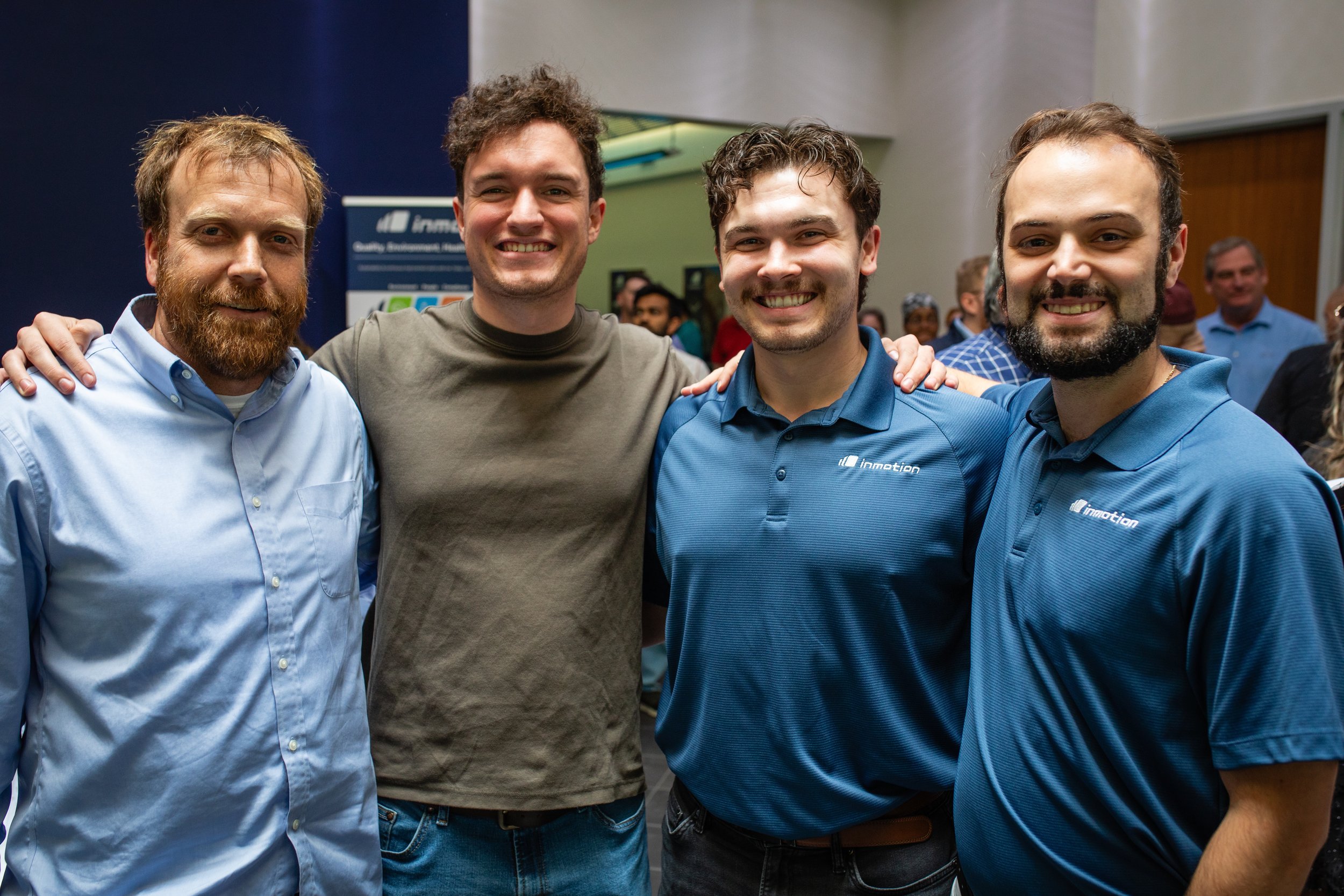 Four men standing together at an indoor event, smiling for the camera. Two of them are wearing blue polo shirts with a company logo, one in a gray T-shirt, and the fourth in a light blue button-up shirt. The background shows other people and a banner
