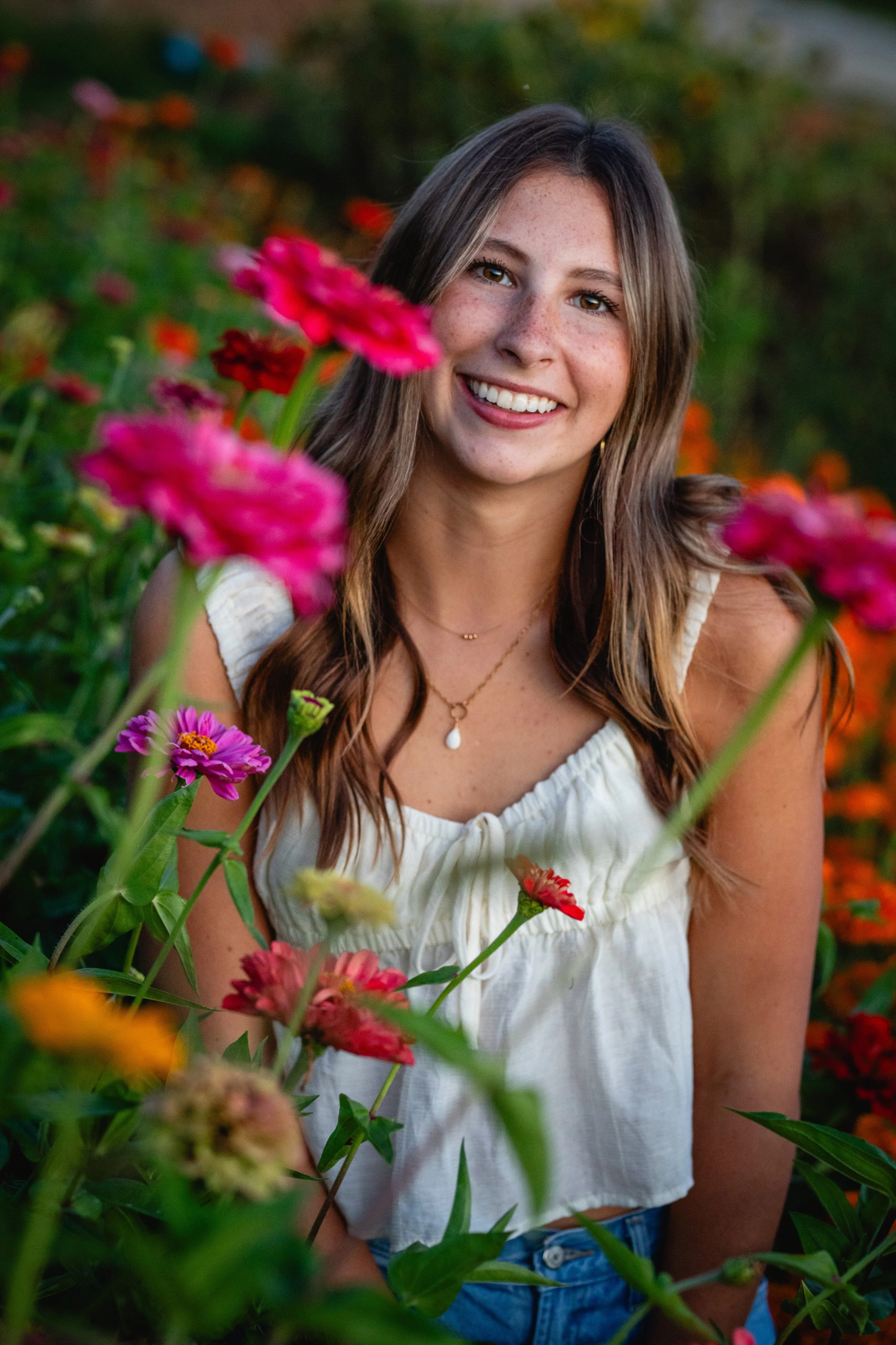 A young woman smiling in a field of colorful flowers, wearing a white sleeveless top and a necklace.