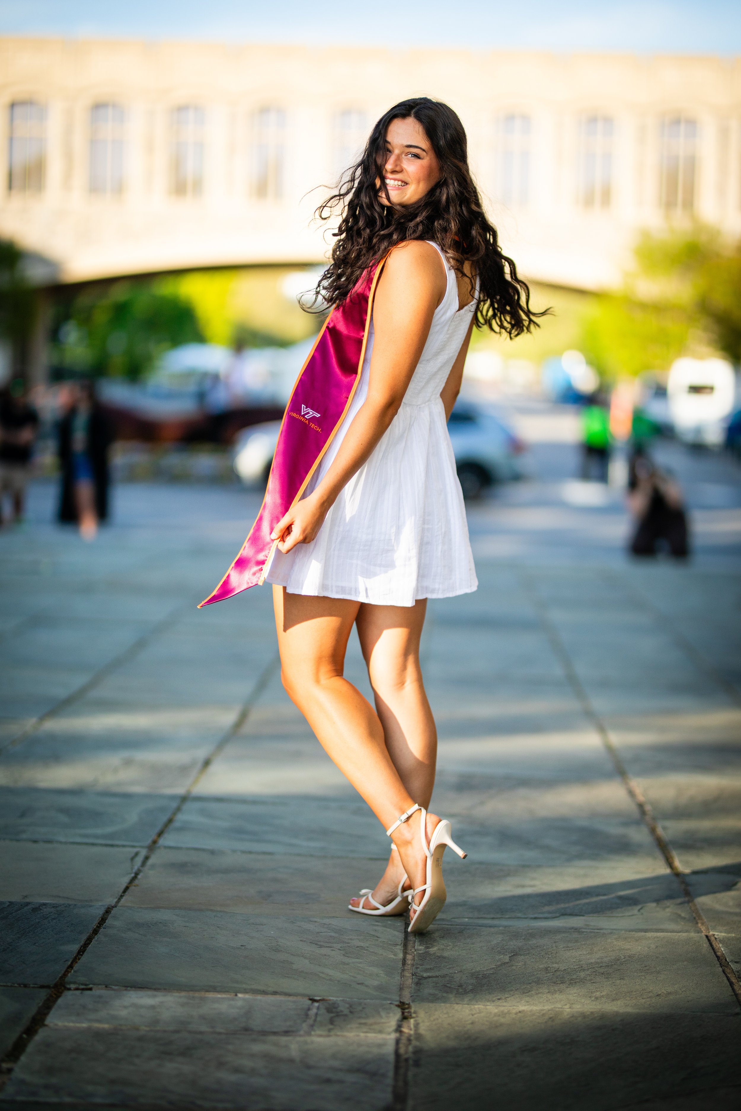 A young woman with curly dark hair wearing a white dress and high heels is smiling and posing outdoors during daylight. She has a maroon graduation sash that reads 'VIRGINA TECH' and 'VT' with a gold trim. The background shows a stone walkway, some b