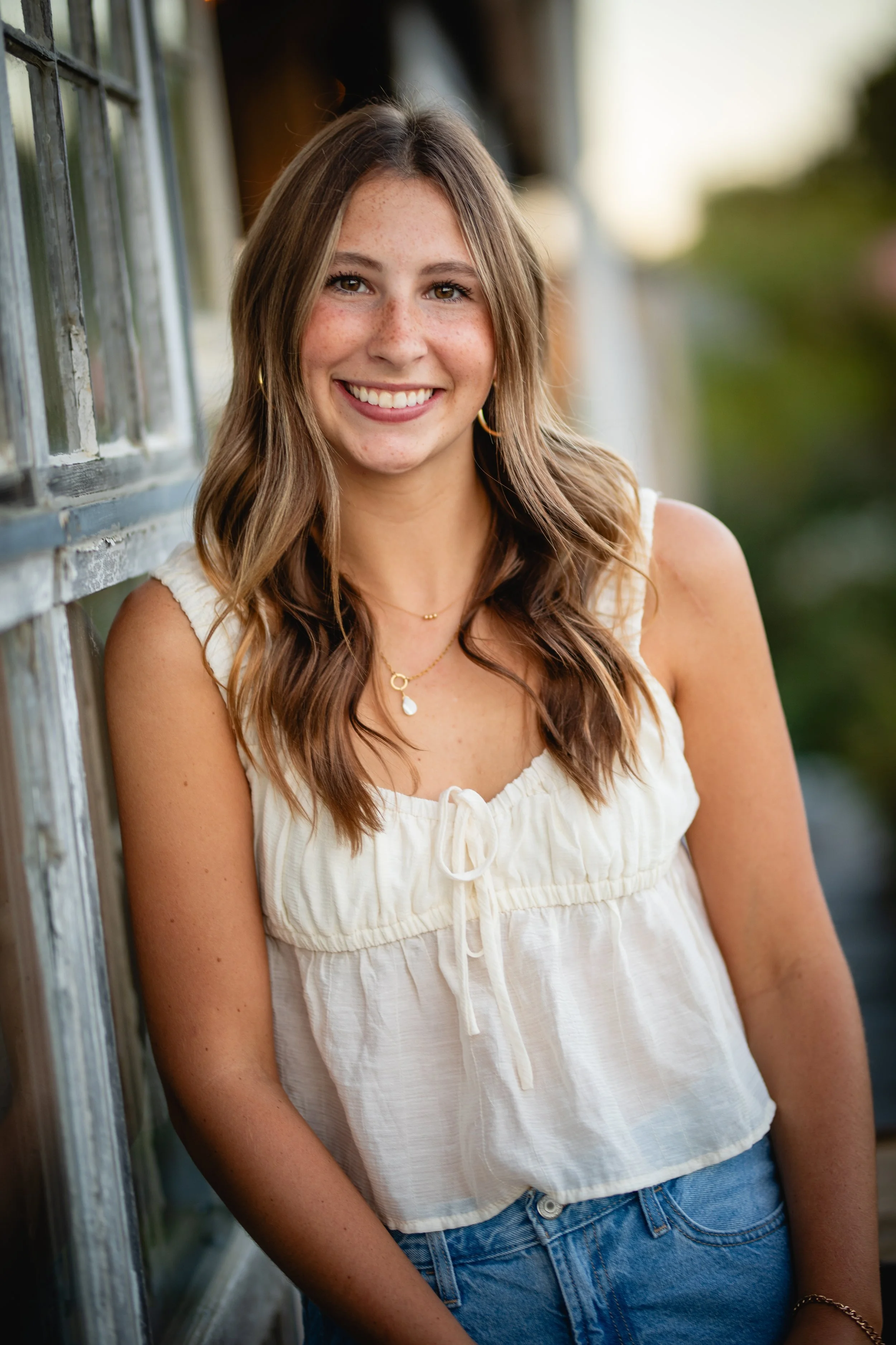 A young woman with wavy brown hair, wearing a cream-colored sleeveless top and blue jeans, smiling and leaning against a weathered wooden window frame outdoors during daytime.