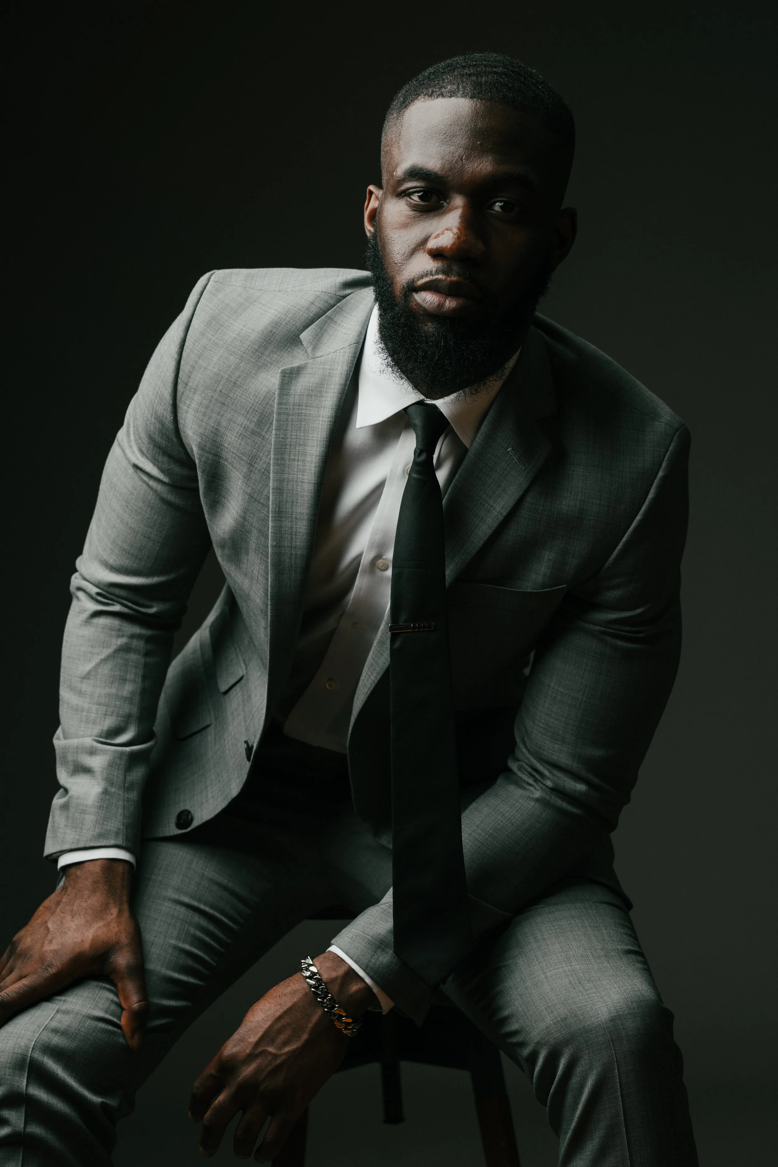 A man wearing a gray suit, white shirt, and black tie, sitting on a stool against a dark background.