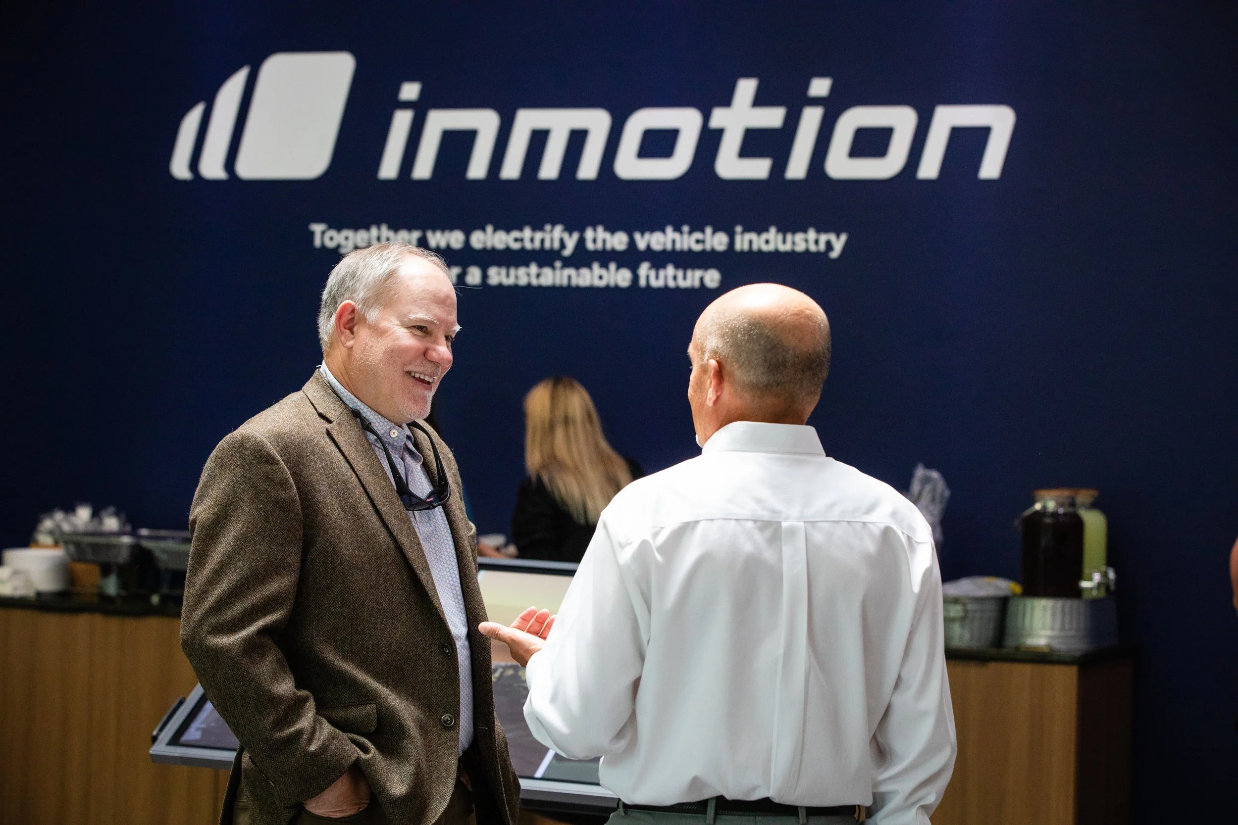 Two men in conversation at an event, with a blue wall behind them displaying the logo and slogan of 'imotion'.