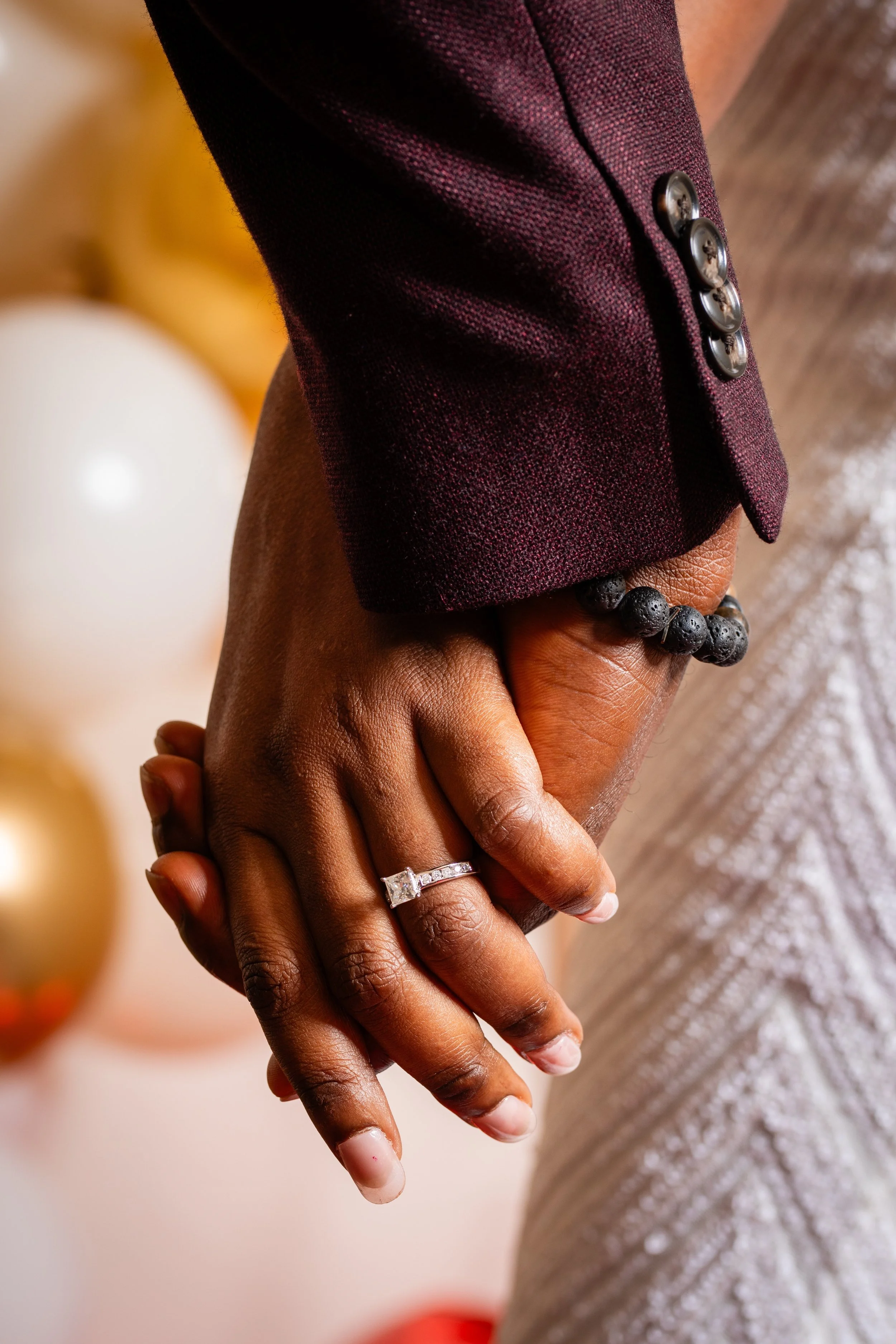 Close-up of a couple holding hands, with the person on the left wearing a ring and a beaded bracelet, dressed in a maroon suit jacket, during a celebration or event with balloons in the background.