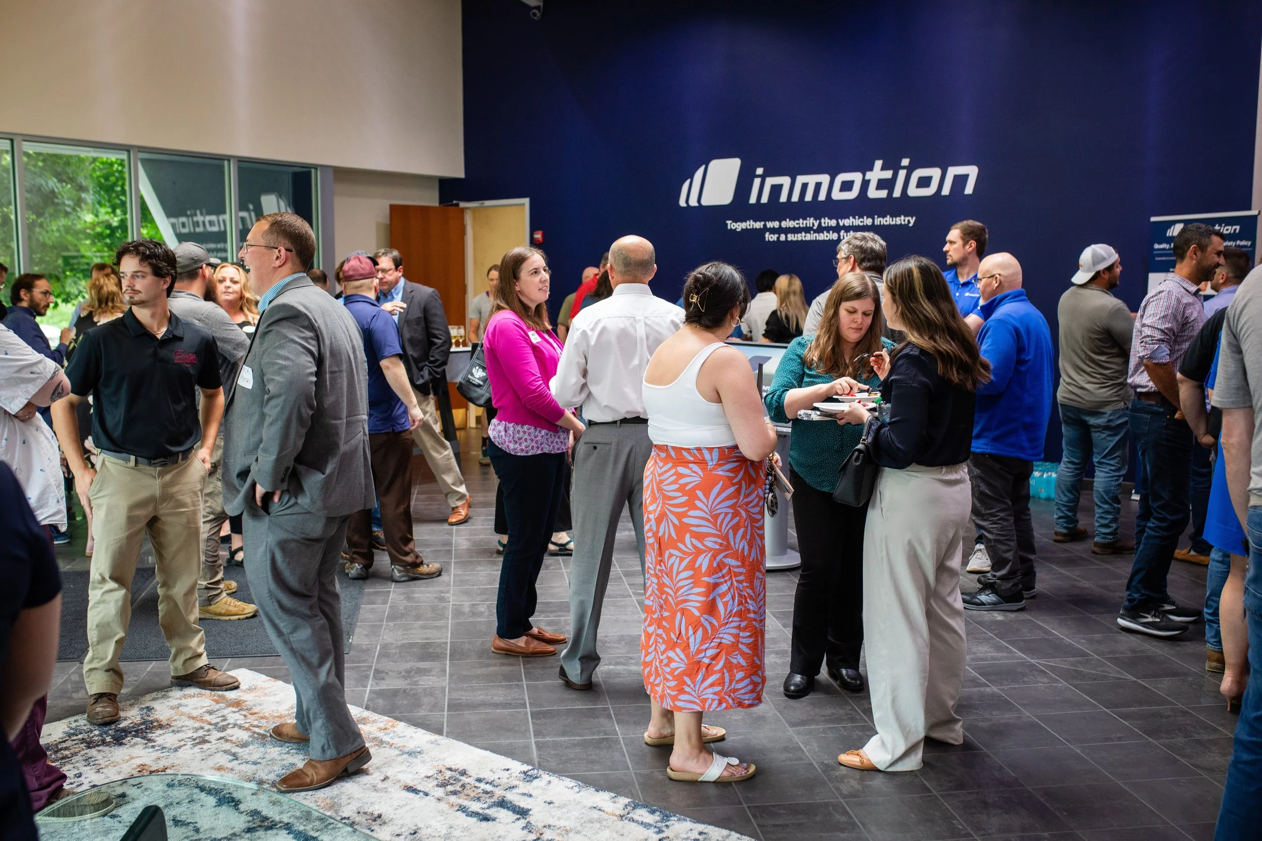 A busy conference or trade show with people engaging in conversations, standing in line, or browsing booths. The backdrop features the InMotio logo and slogan about electrifying the vehicle industry for a sustainable future.