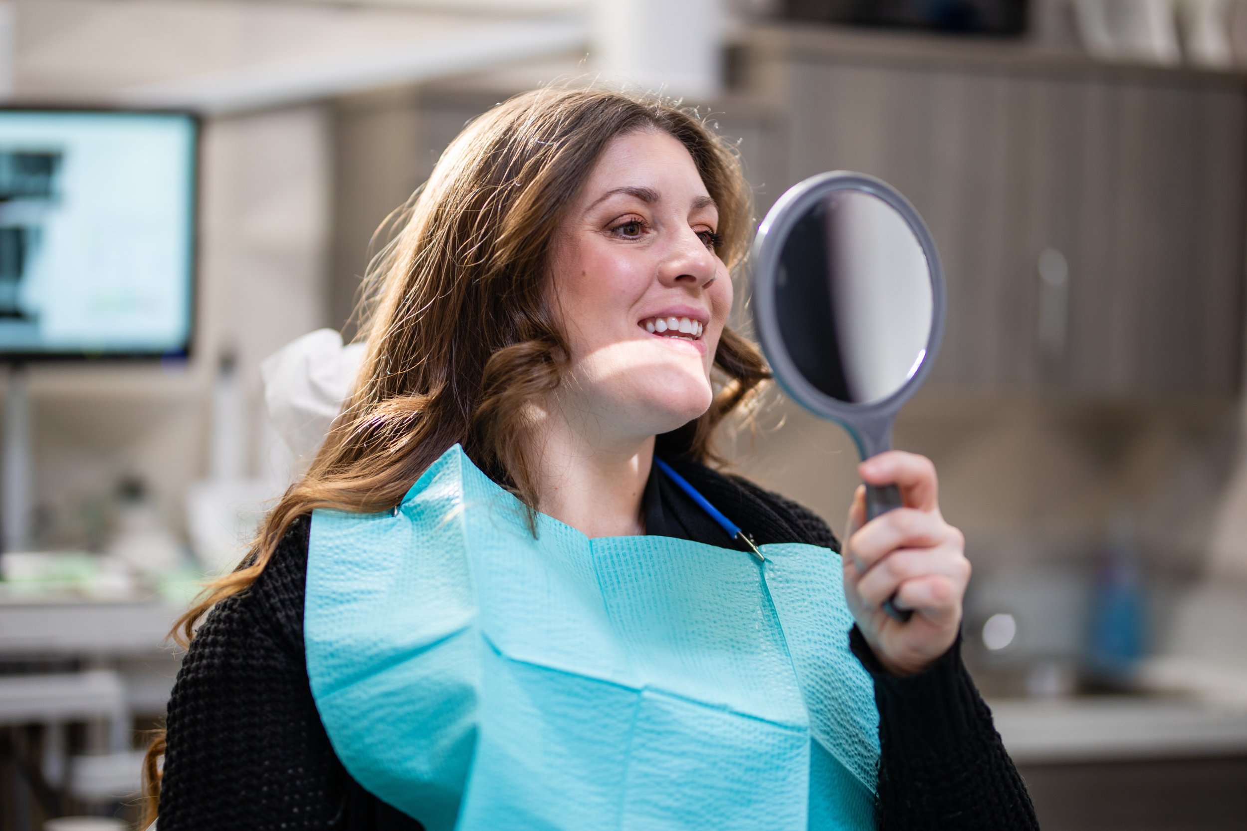 A woman in a dental clinic holding a mirror and smiling, wearing a dental bib and black sweater.