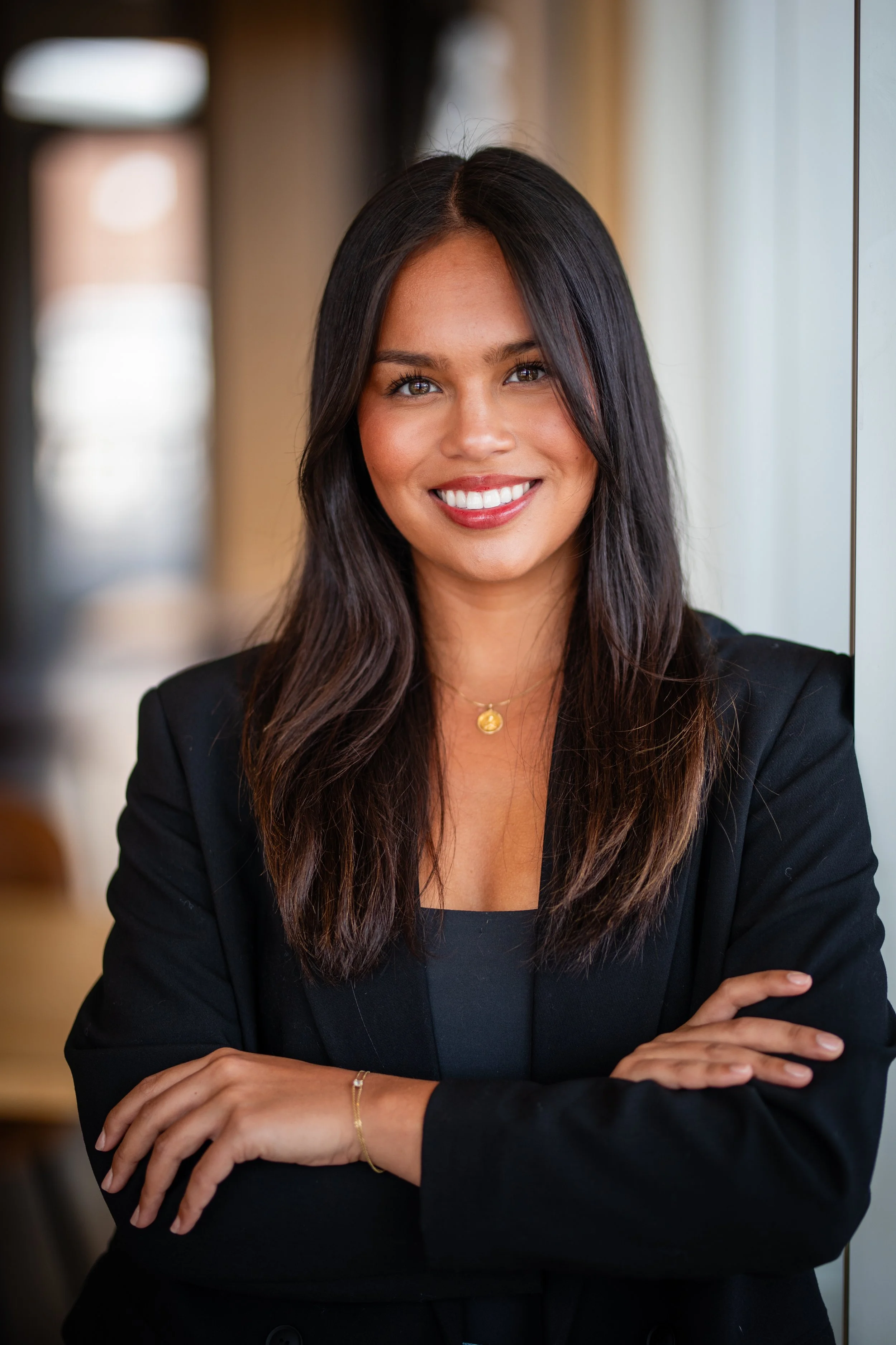 A woman with long dark hair smiling, wearing a black blazer and gold jewelry, standing indoors near a window with her arms crossed.