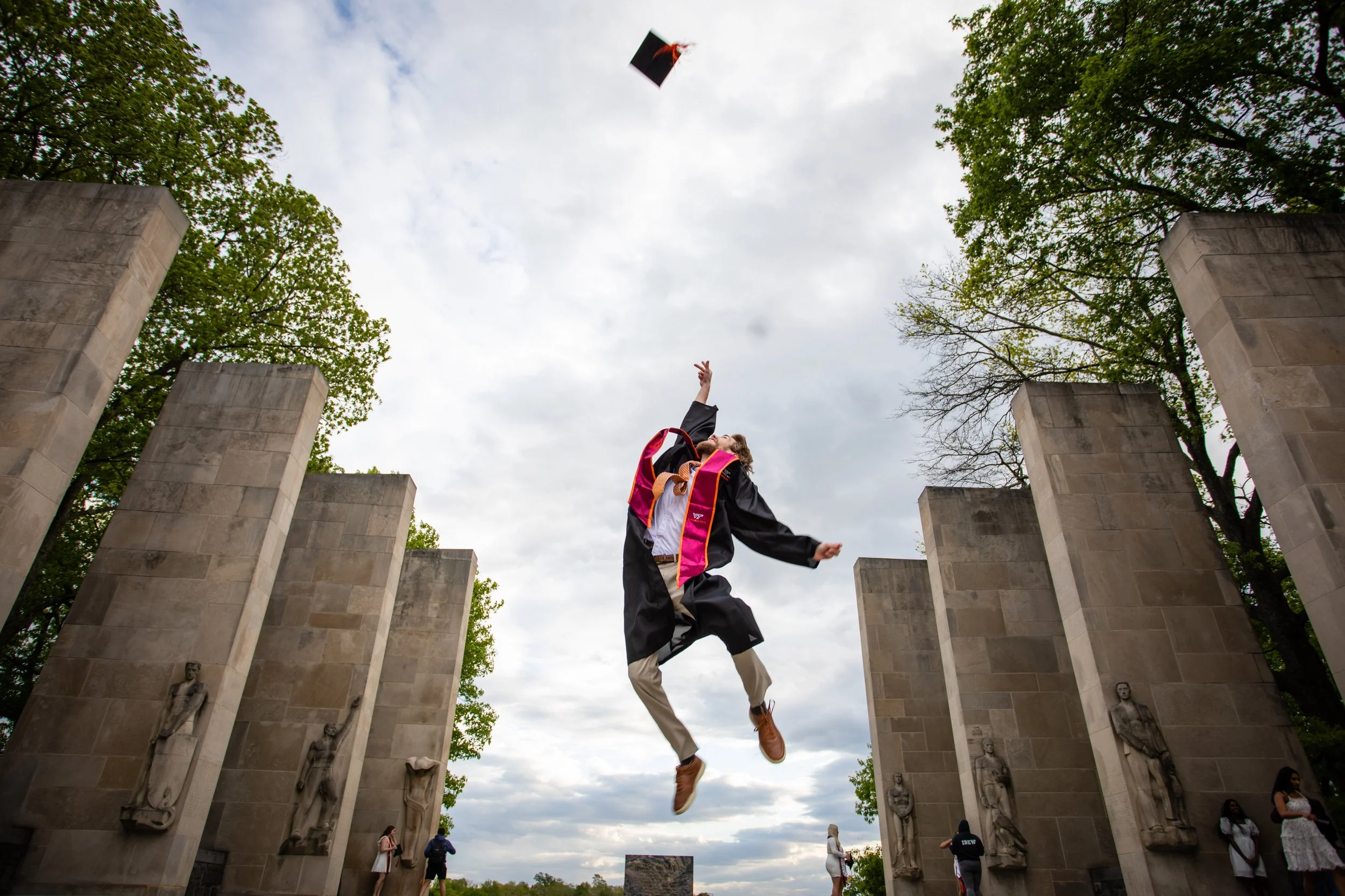 Graduate in black cap and gown jumping in the air during graduation ceremony at a memorial site, with other graduates and visitors in the background, under cloudy sky.