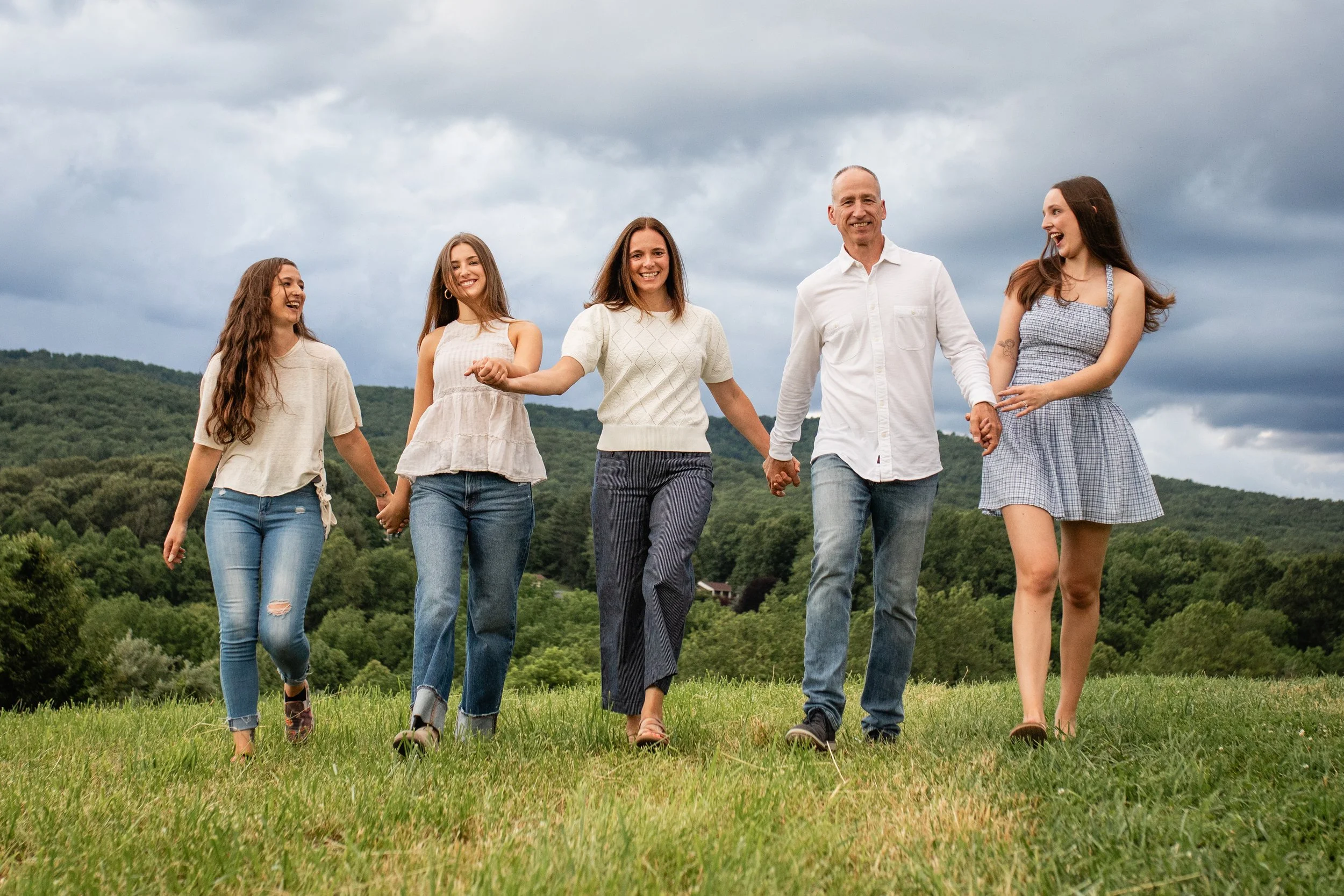 Family of six walking hand-in-hand outdoors in a green field with hills and cloudy sky in the background, smiling and laughing.