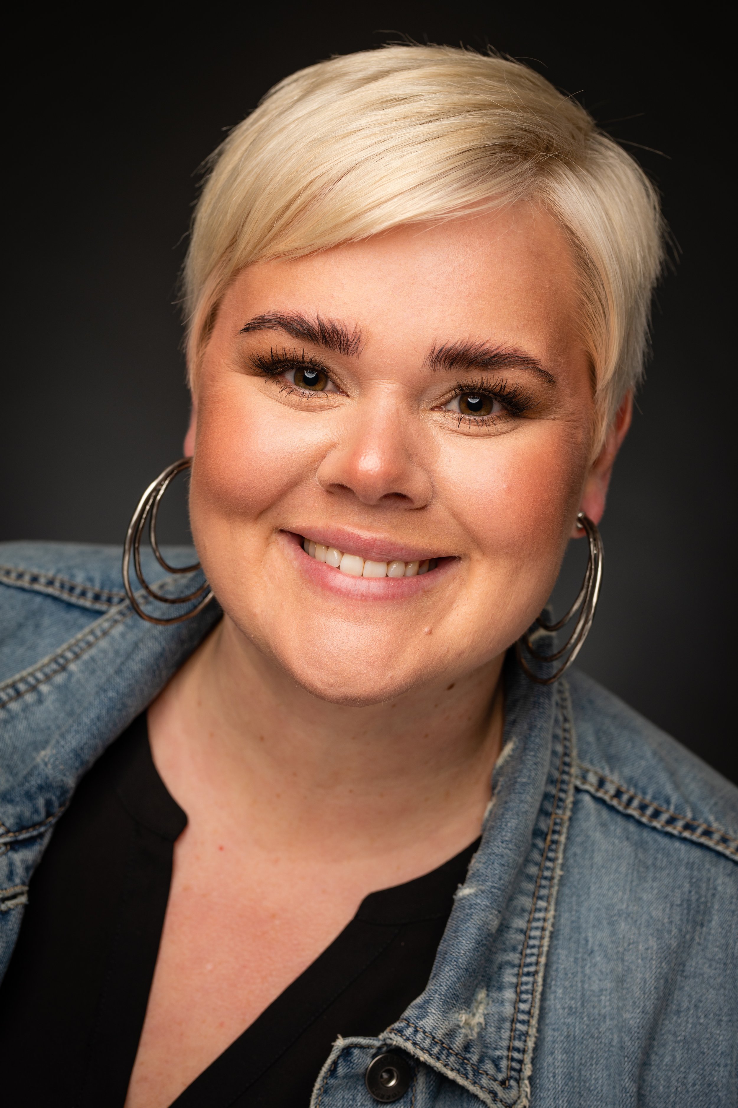 Close-up portrait of a smiling woman with short blonde hair, wearing hoop earrings, a black top, and a denim jacket, against a dark background.