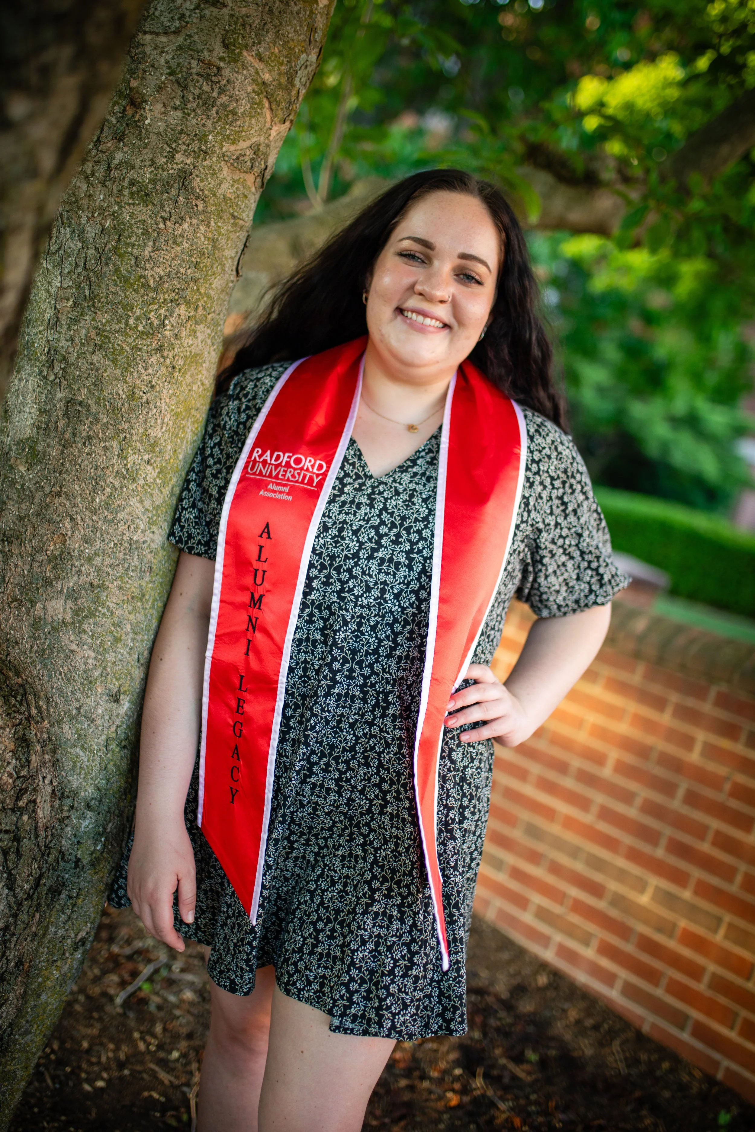 A young woman in a black floral dress with a red graduation stole from Radford University, smiling and standing outdoors near a tree and brick wall.