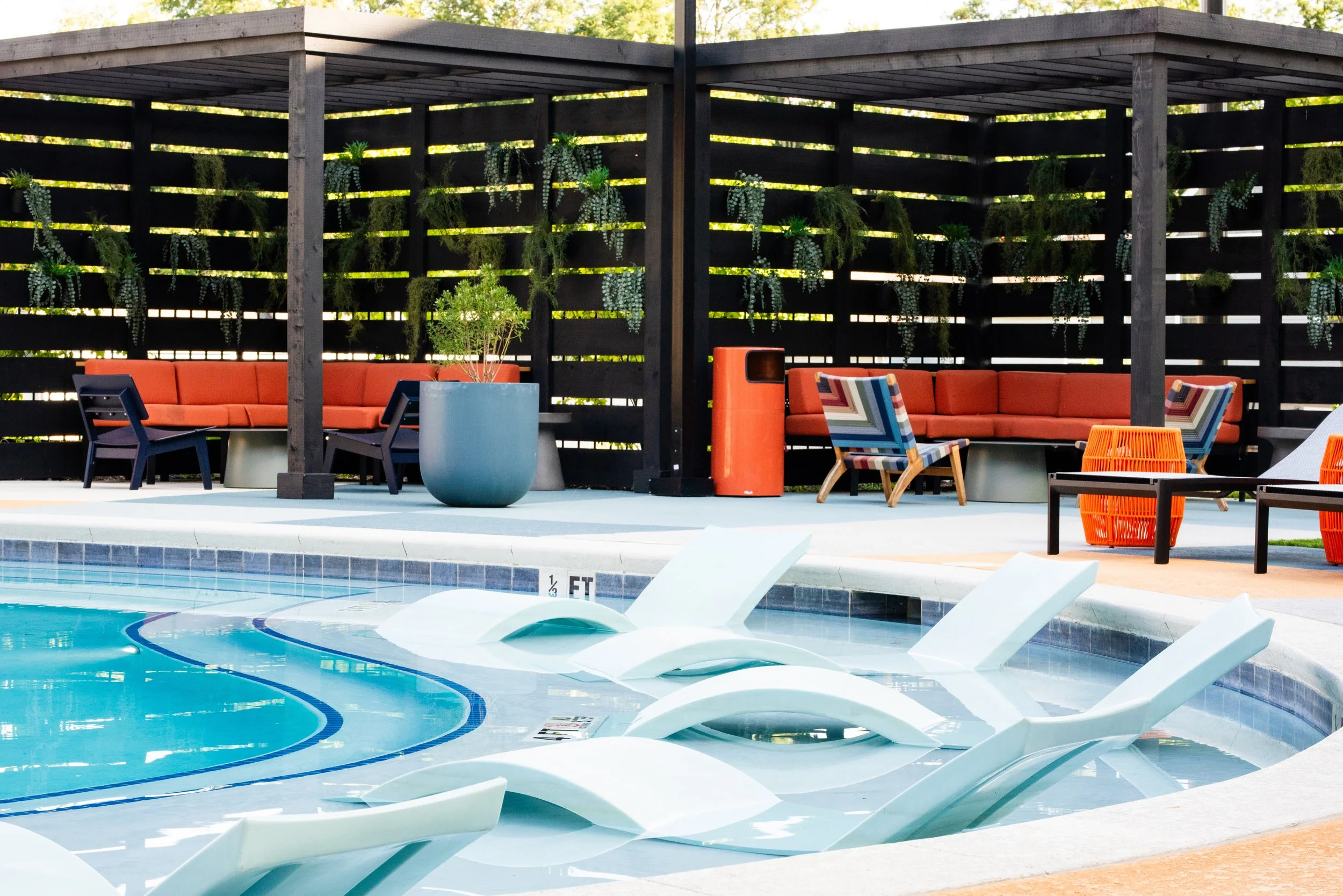 An outdoor pool area with white pool loungers partially submerged in the water, surrounded by a deck with modern chairs, benches, and potted plants under a wooden pergola.