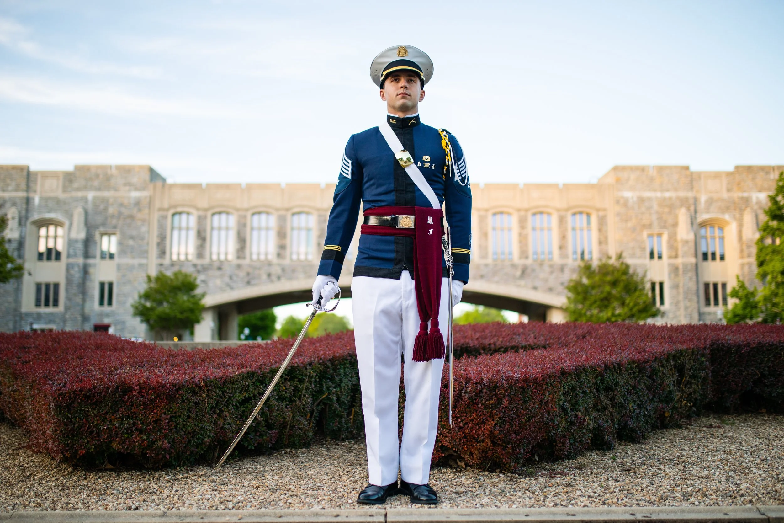 A young man dressed in a traditional military cadet uniform standing outdoors in front of a historic stone building with an archway, surrounded by neatly trimmed bushes and trees.
