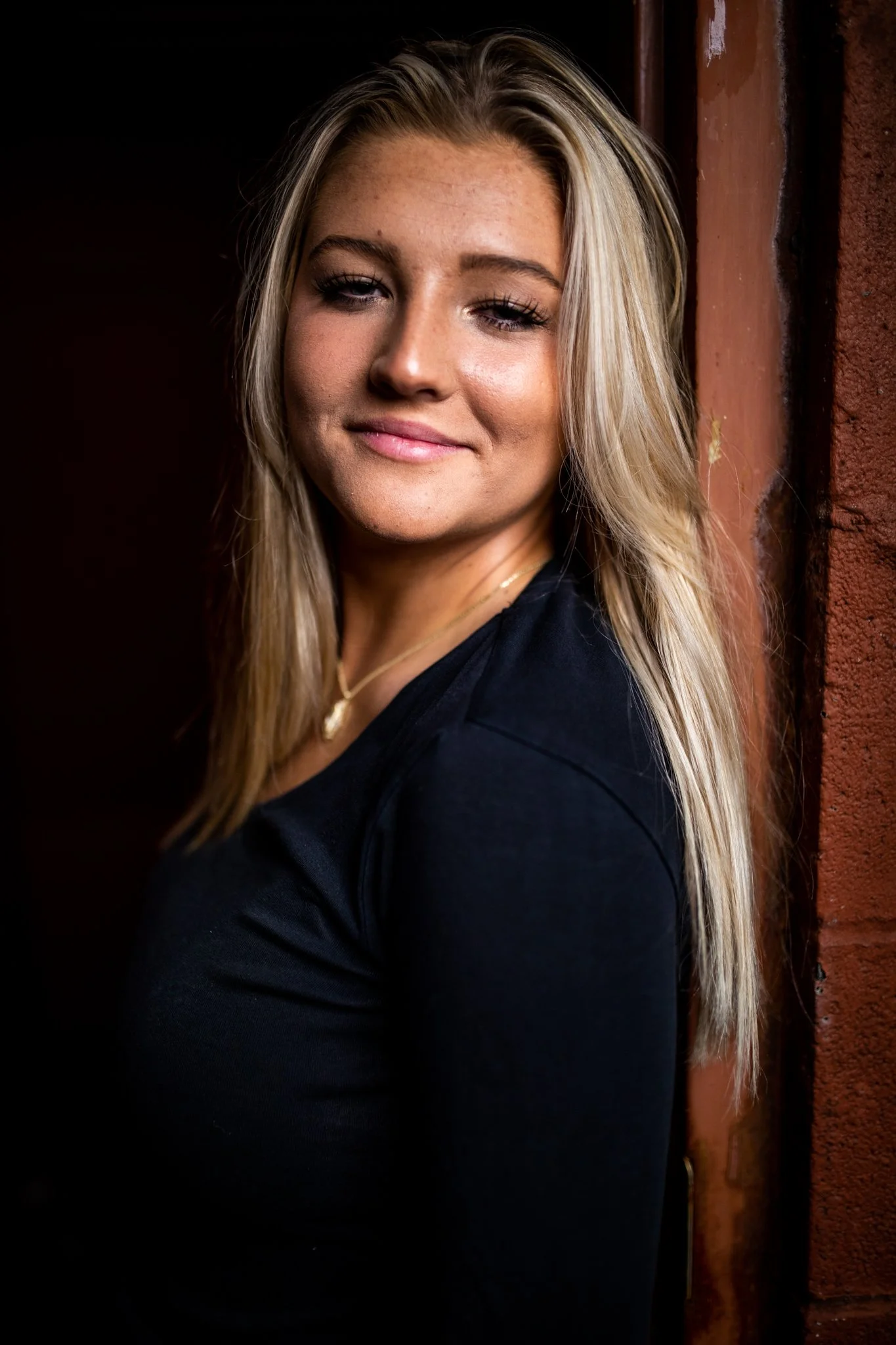 A young woman with long blonde hair leaning against a brick wall, wearing a black top and a gold necklace, smiling softly.