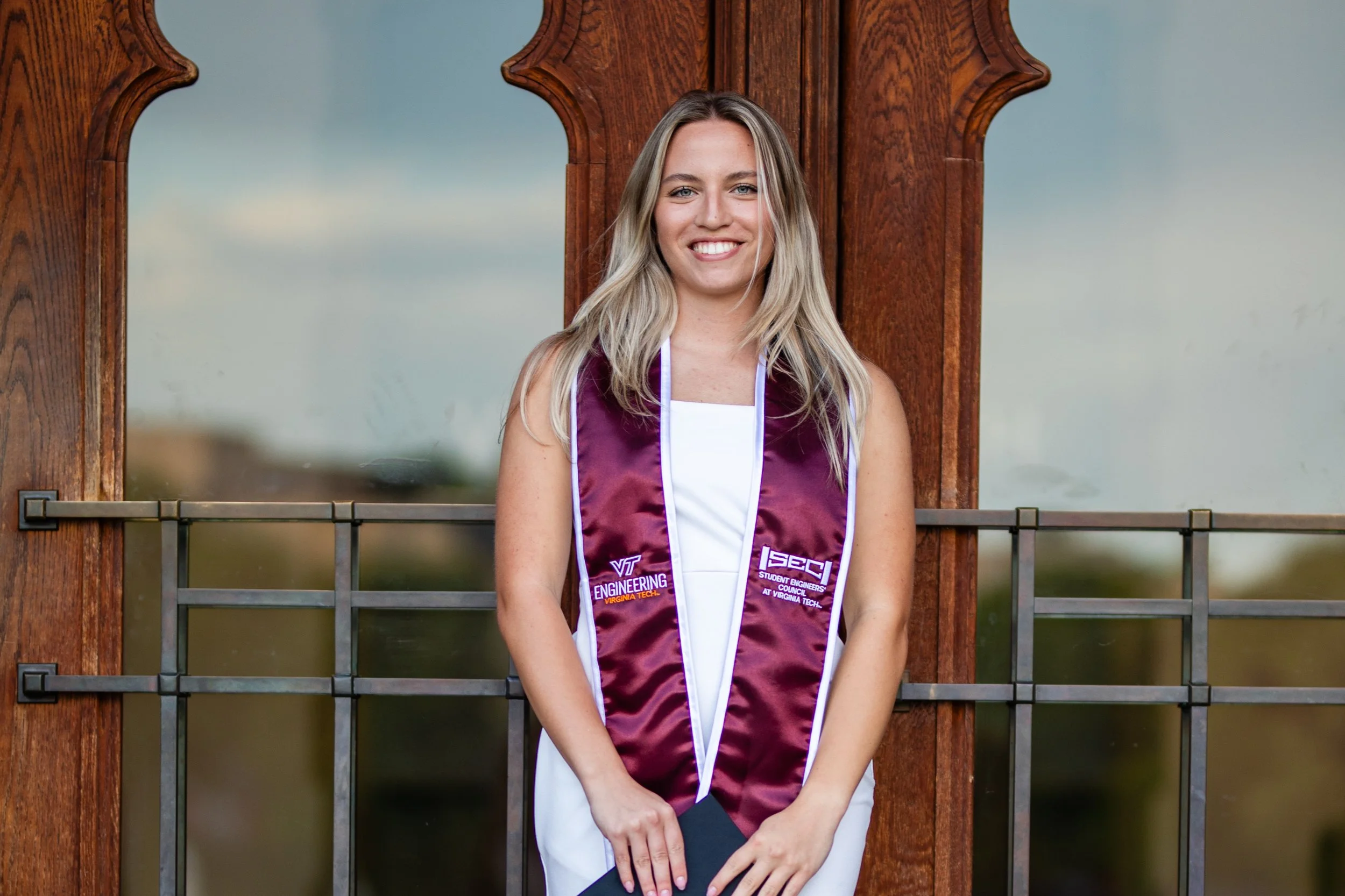 Young woman in graduation gown standing outdoors in front of a wooden and glass structure, smiling at the camera.