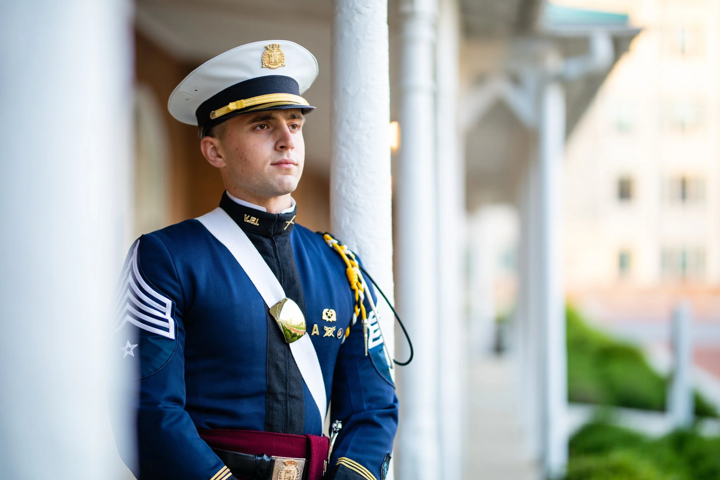 Young military cadet in blue uniform and white cap standing outdoors next to white columns.