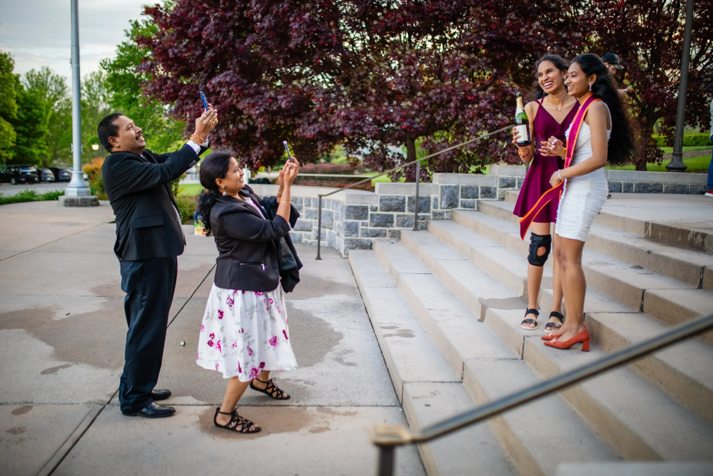 A group of people celebrating outdoors on a staircase with trees in the background, some holding drinks and capturing moments with their phones, dressed in semi-formal attire.