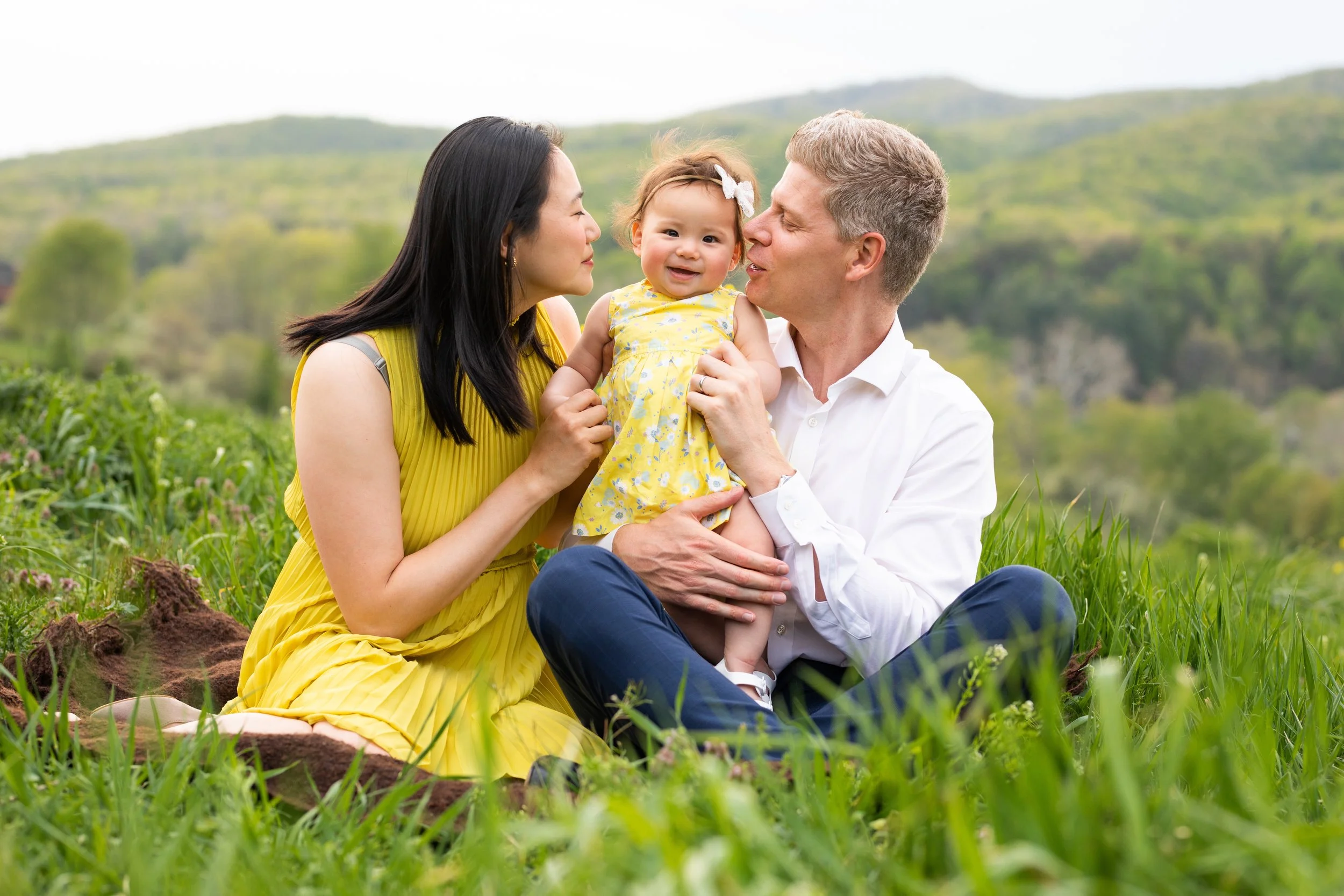 A family sitting on grass in a field with a scenic green hillside in the background. The family includes a woman in a yellow dress, a man in a white shirt, and a young girl in a yellow dress, all smiling and interacting affectionately.
