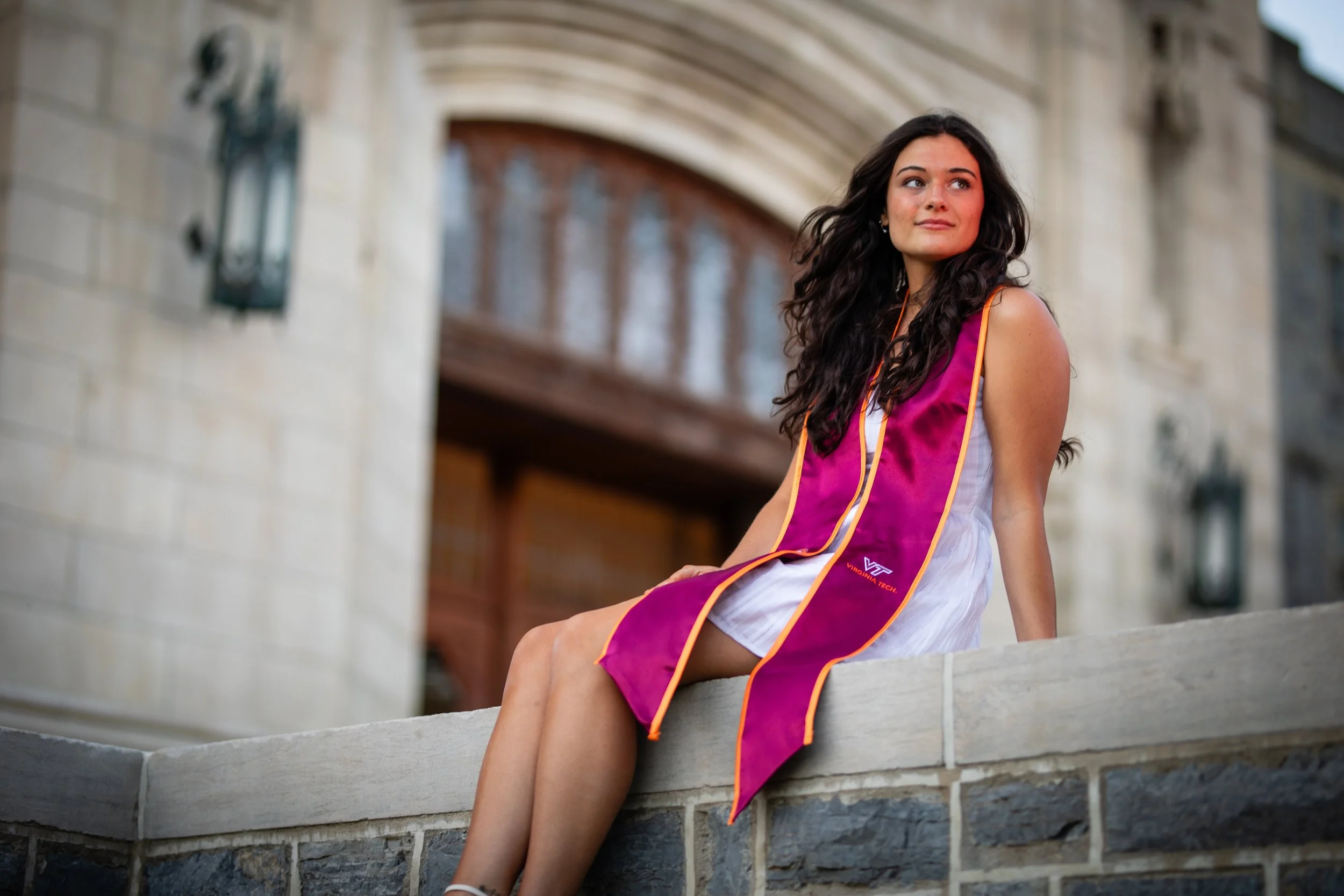 A young woman with long dark curly hair sitting on a stone ledge outside a building, wearing a graduation stole over her white dress, looking to the side with a thoughtful expression.