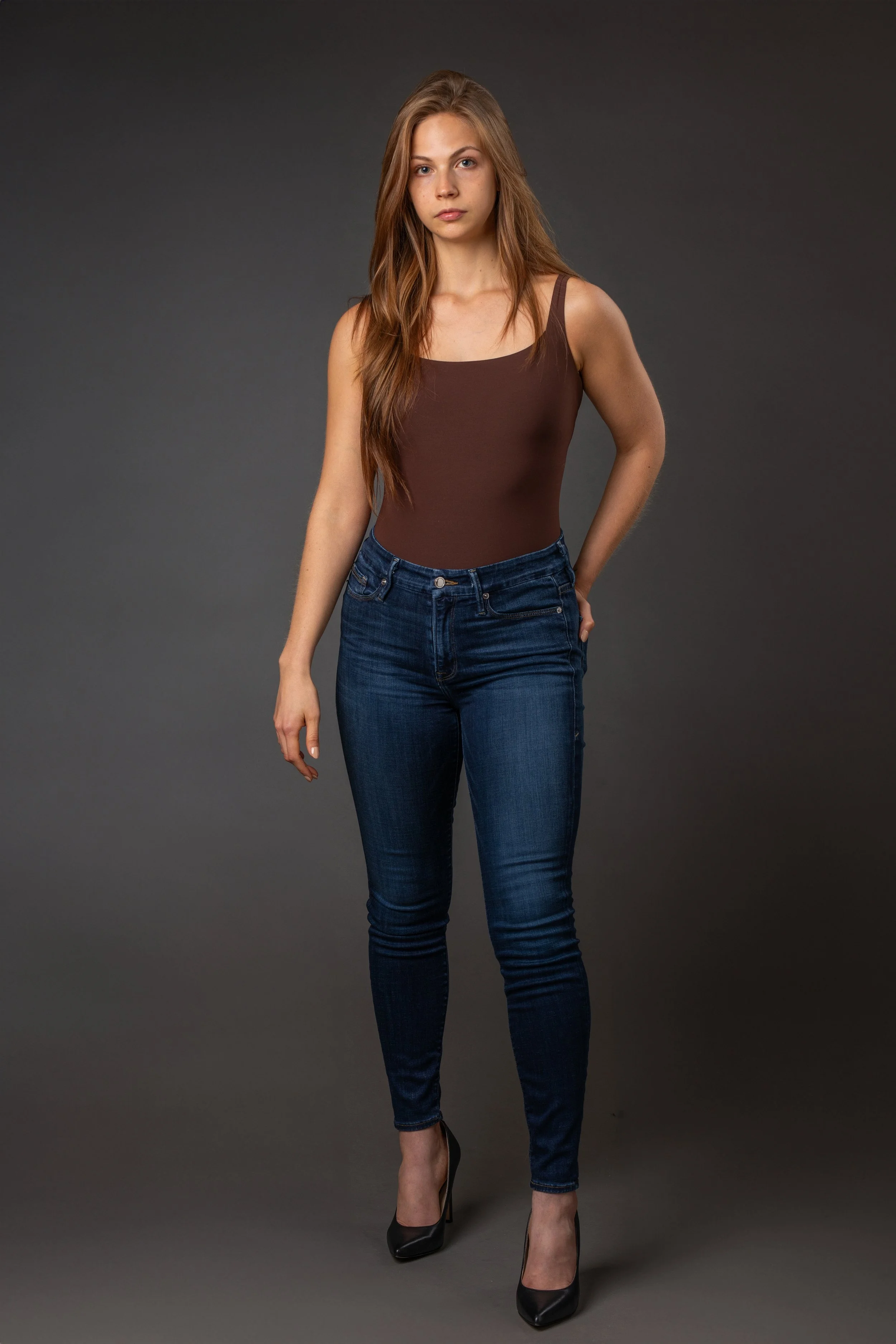 A young woman with long, wavy brown hair stands against a plain gray background. She is wearing a fitted brown tank top, dark blue jeans, and black high heels.