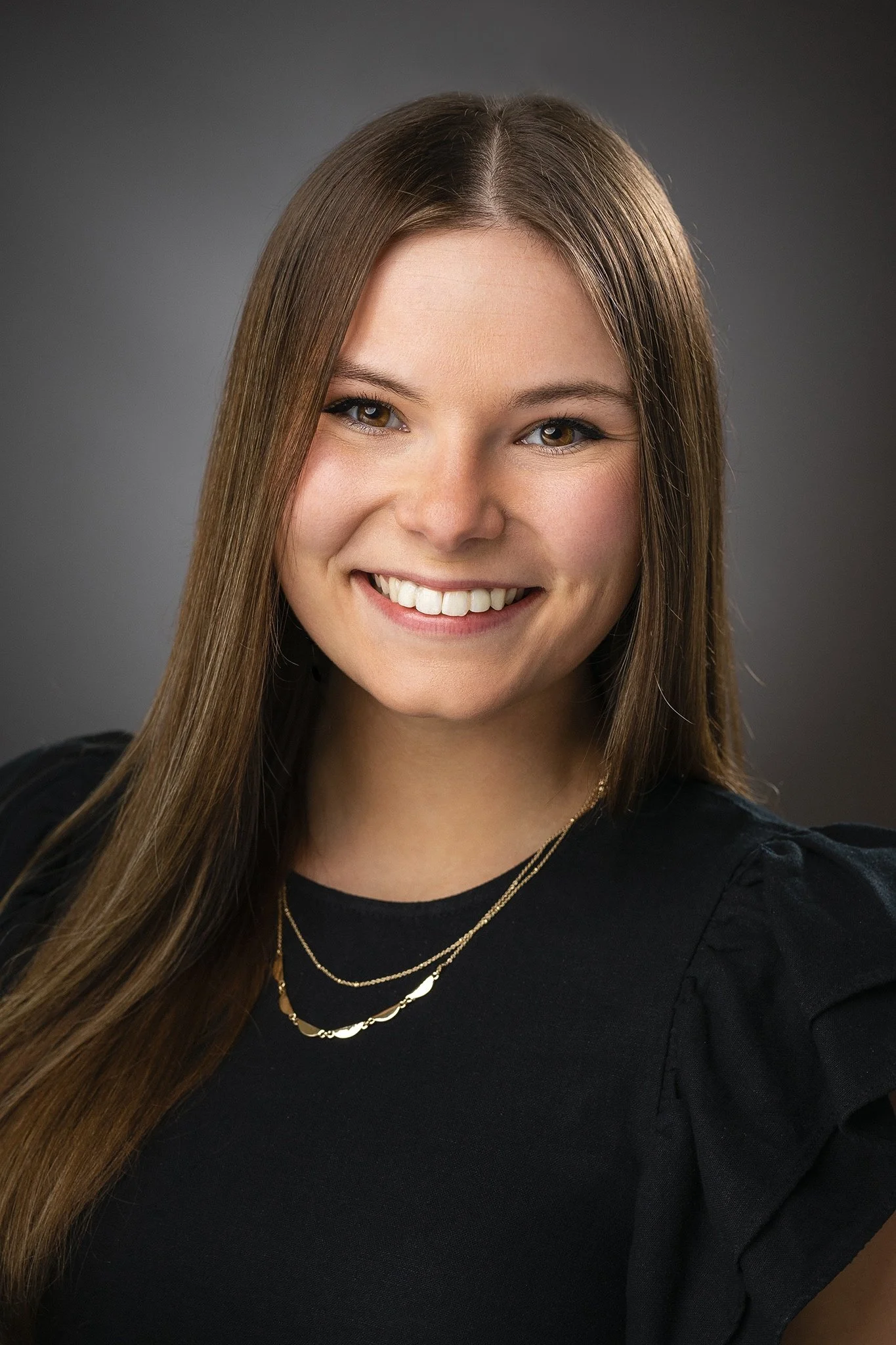 Headshot of a young woman with long brown hair, smiling, wearing a black top and layered necklaces, with a dark gray background.