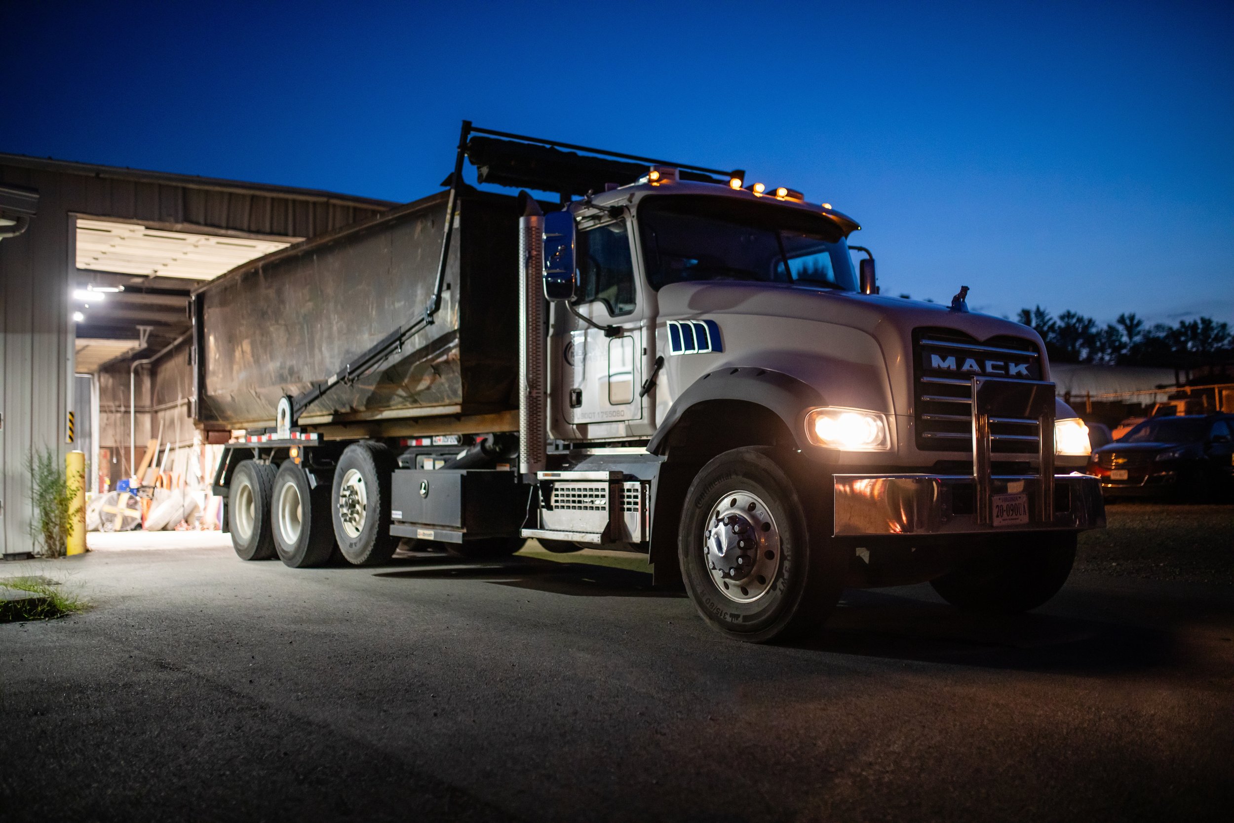 A Mack dump truck with its headlights on parked outside a building at dusk, with a dark blue sky and trees in the background.