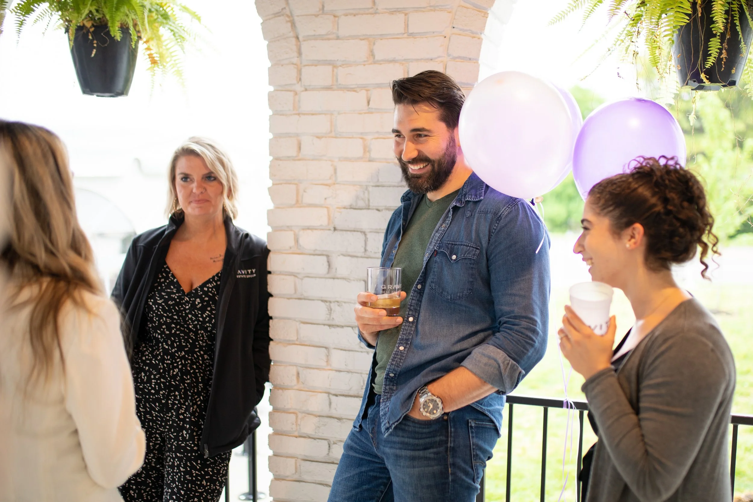 A group of people socializing at a party, with a man holding a drink, smiling, and women holding cups, with balloons in the background.