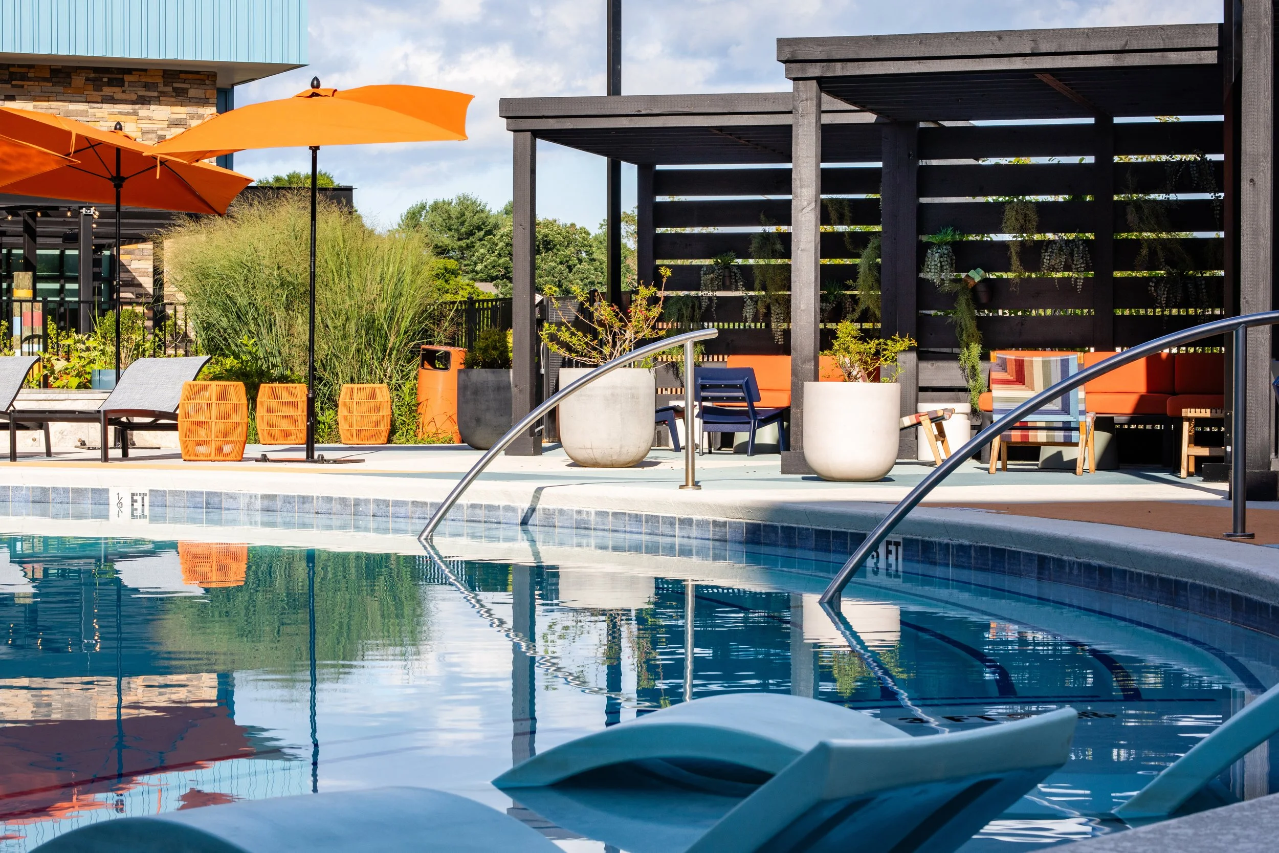 An outdoor swimming pool area with lounge chairs, umbrellas, and a shaded seating area with orange cushions and potted plants.