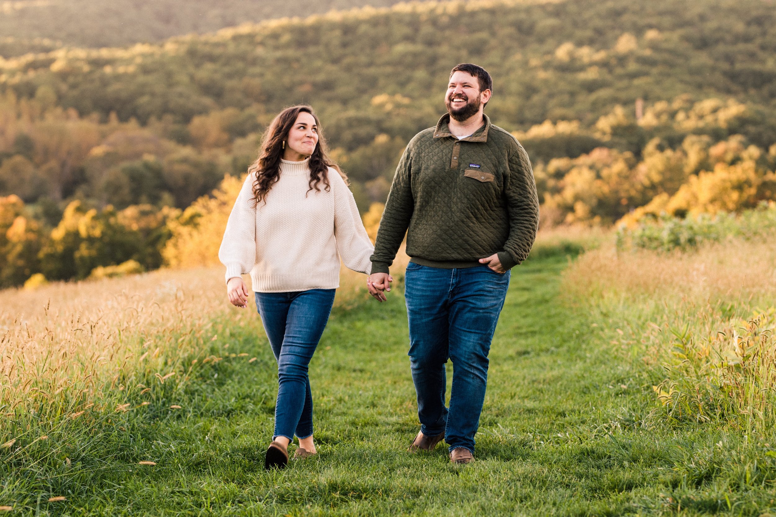 A couple walking hand in hand through a grassy field in autumn, with trees and hills in the background.